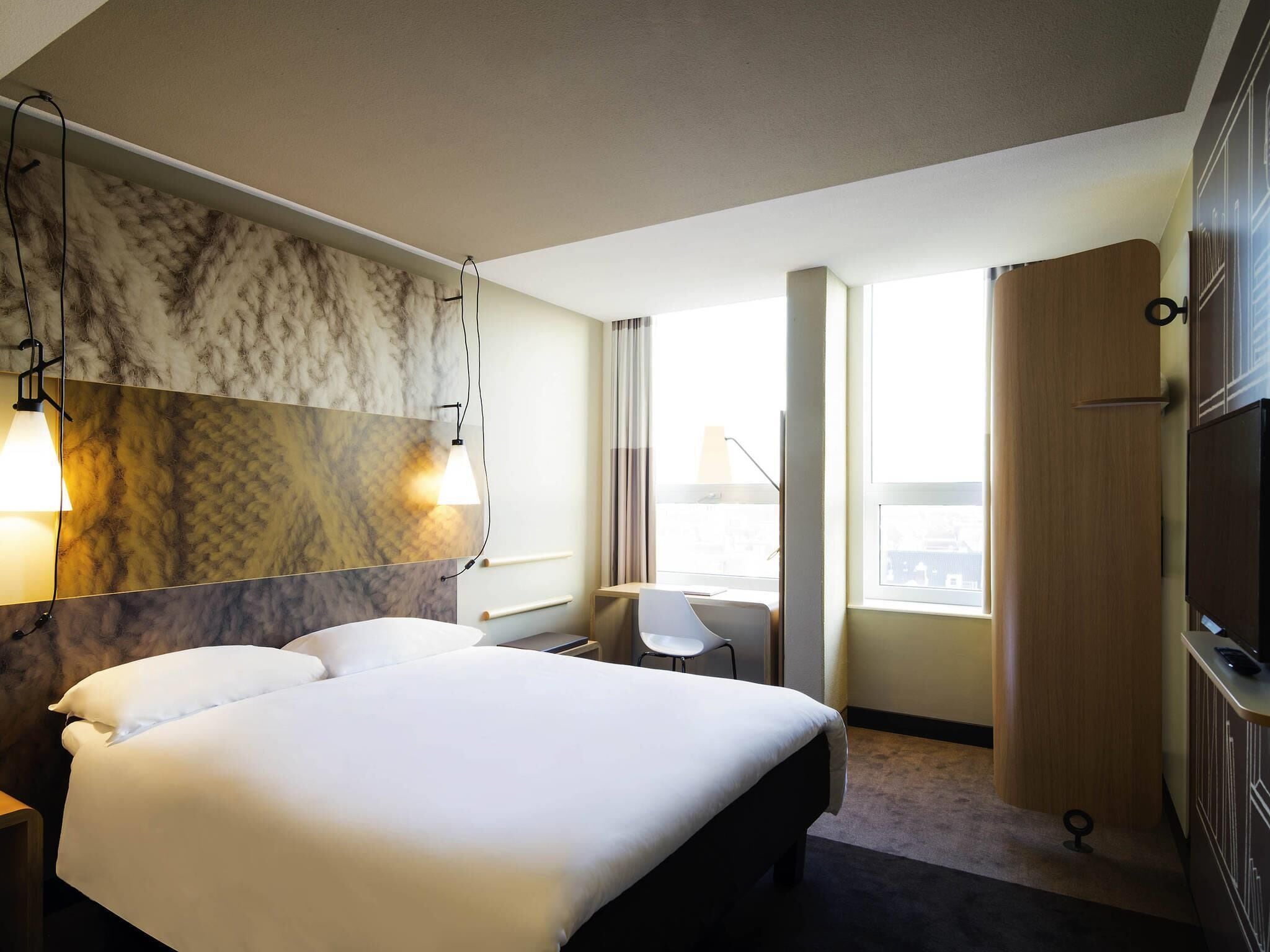 A hotel room with a large bed, a desk, a chair, a wardrobe, and a TV.
