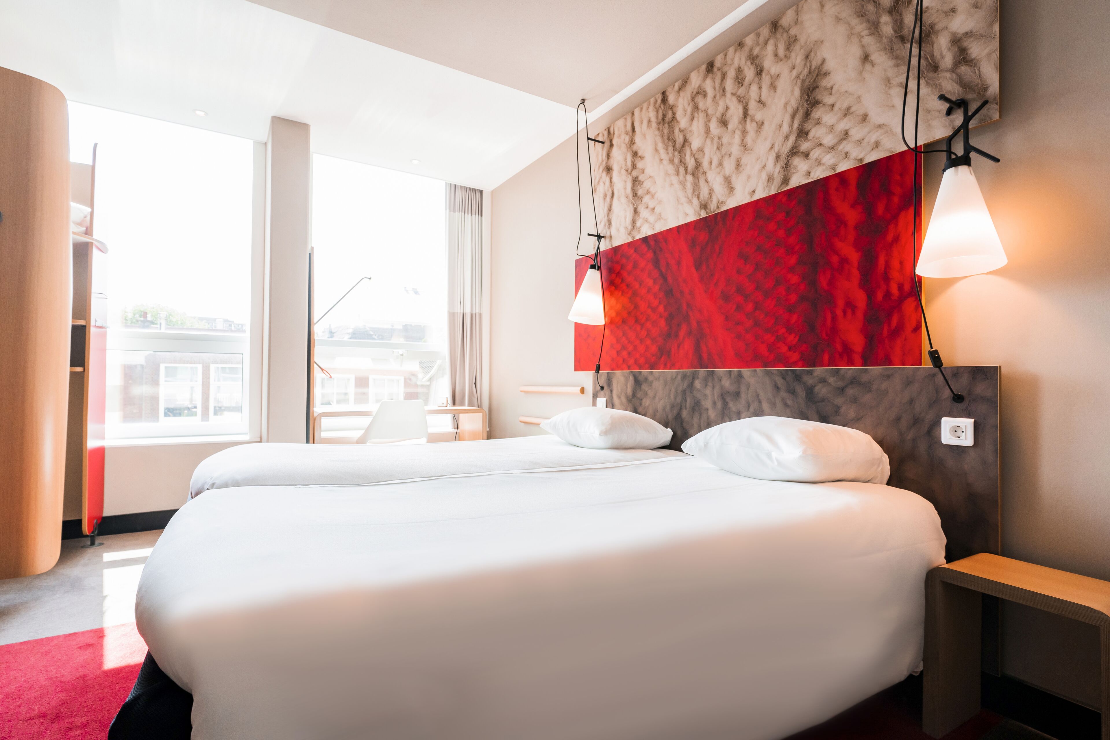 A hotel room with a large bed, a red accent wall, and a view of the city.