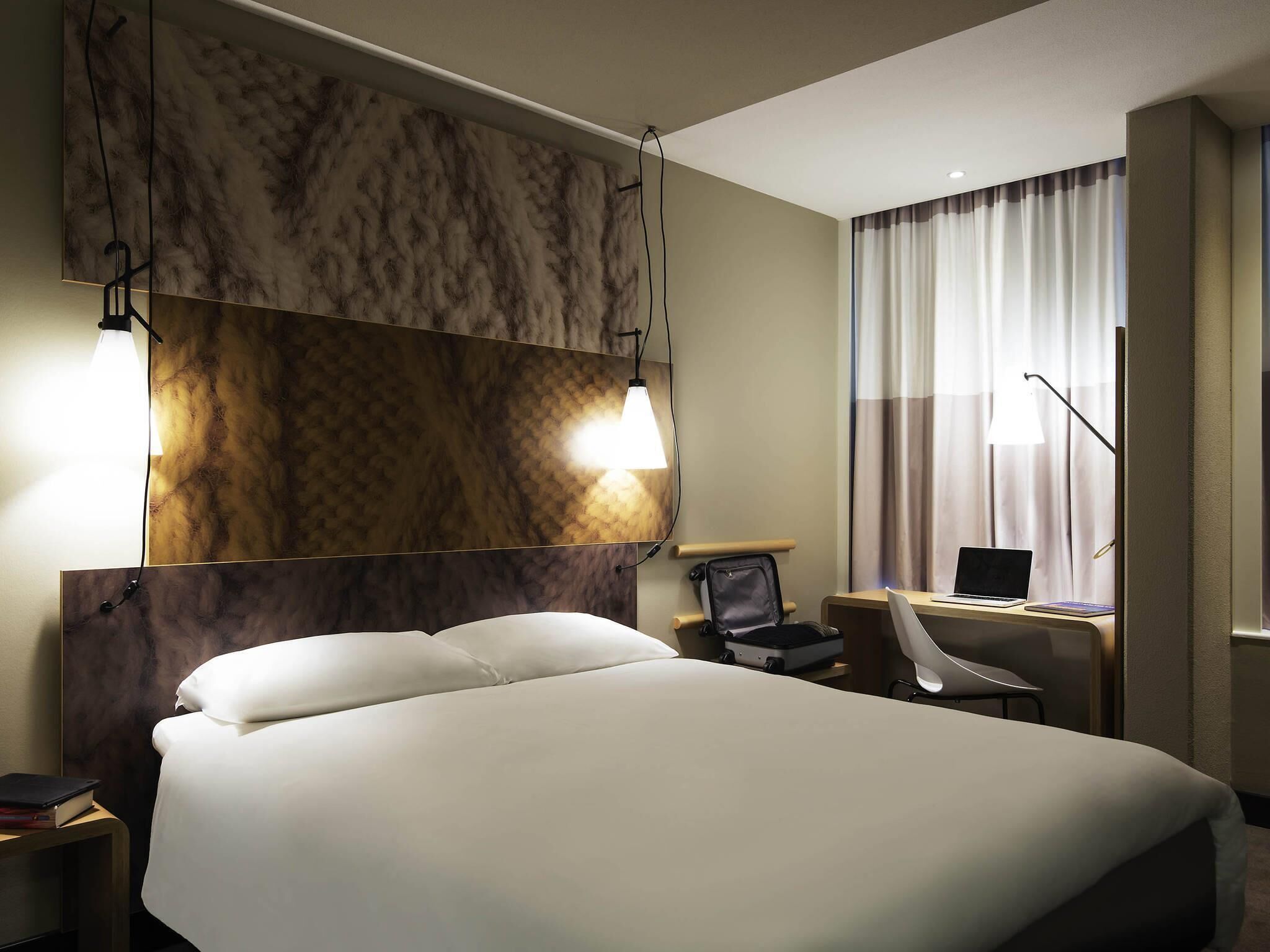 A hotel room with a large bed, a desk with a laptop, a chair, and a view of the outside through curtains.