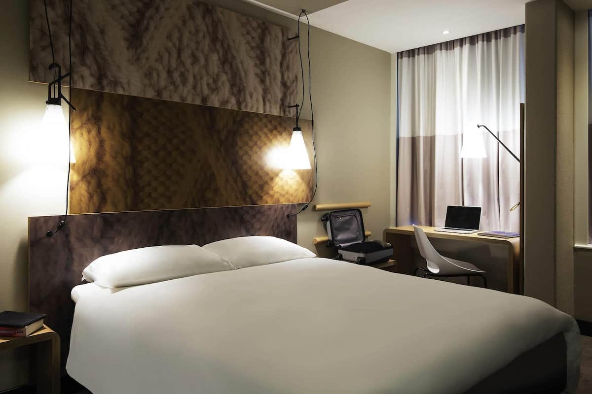 A hotel room with a large bed, a desk with a laptop, a chair, and a view of the outside through curtains.