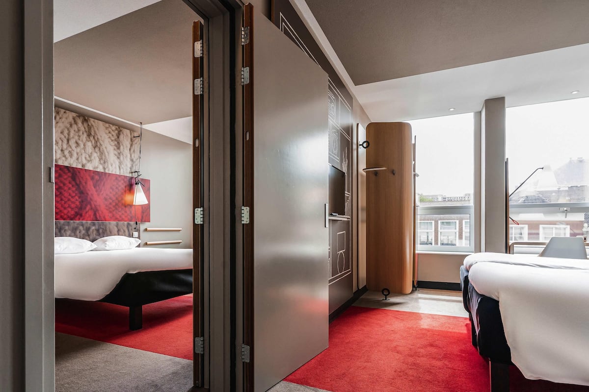 Ein Hotelzimmer mit zwei Betten, einem roten Teppich, einem großen Fenster und Blick auf die Stadt.