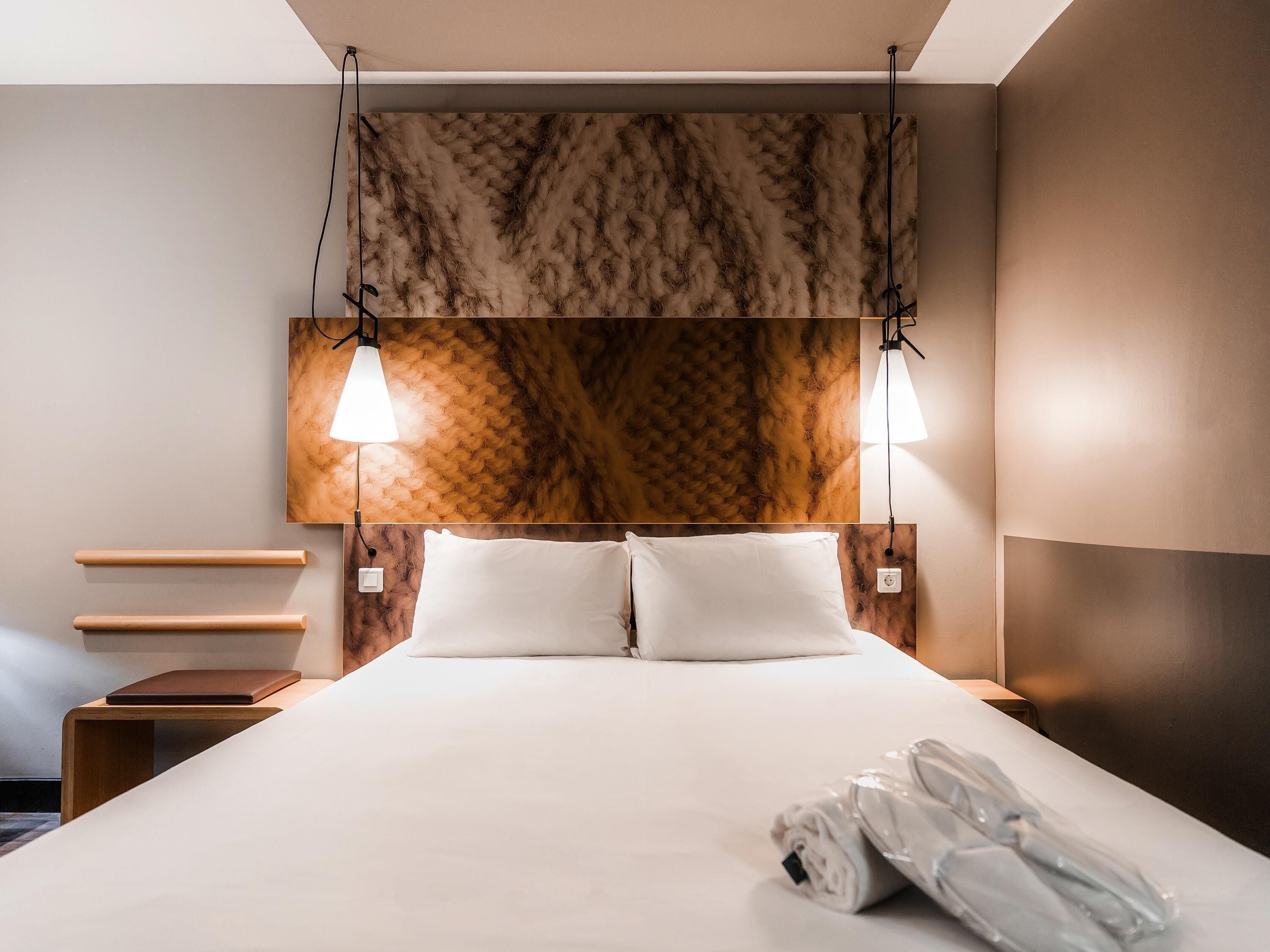 A hotel room with a large bed, two bedside tables, a headboard with a textured pattern, and two wall-mounted lamps.