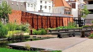 Een voetpad langs de rivier met een houten brug, een bakstenen gebouw en een tuin met diverse planten.
