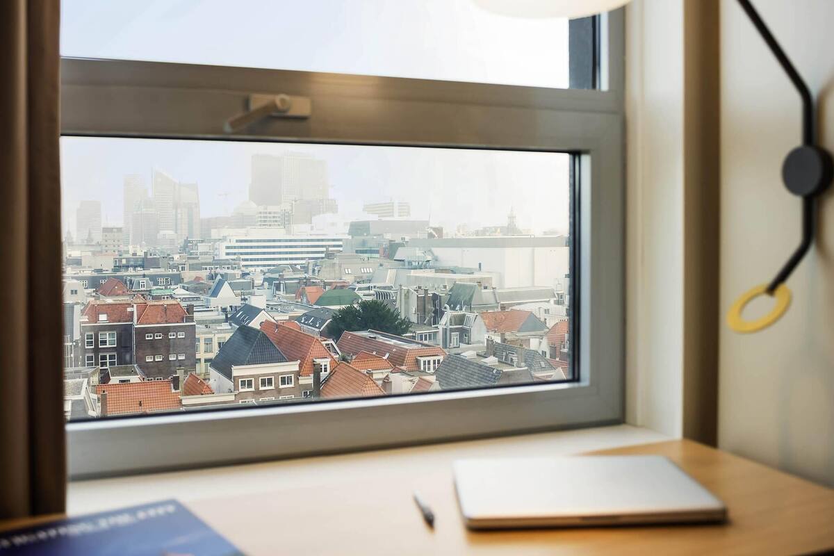 Een raam met uitzicht op een stadsbeeld, een lamp en een notitieboekje op een bureau.