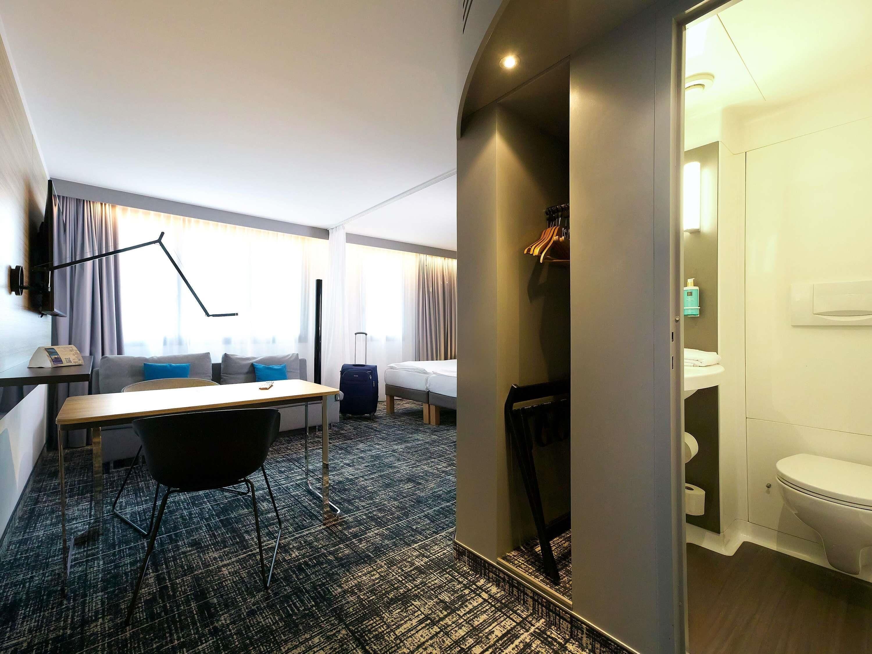 A hotel room with a bed, a desk, a chair, a suitcase, and a bathroom.