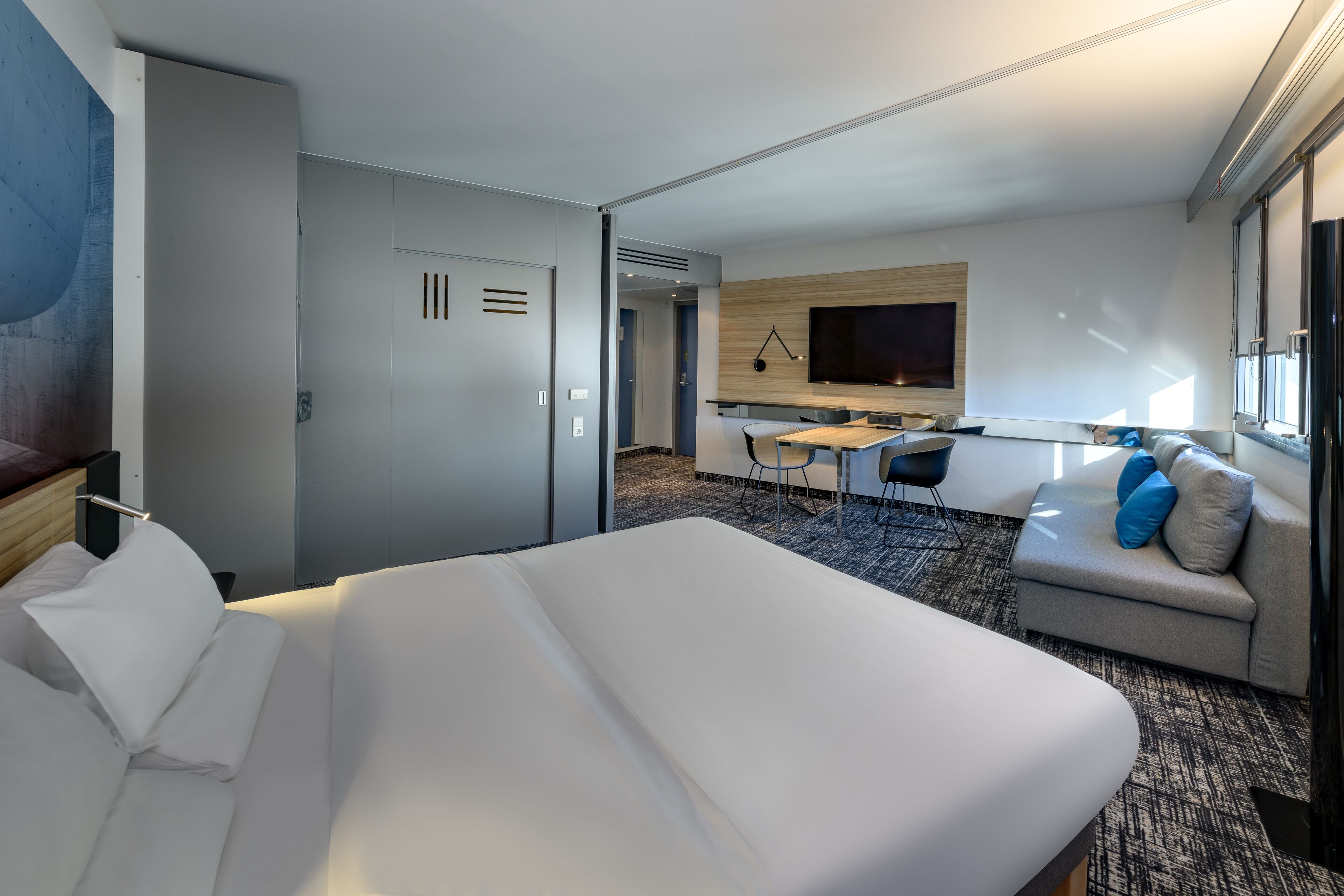 A hotel room with a large bed, a desk with a chair, a sofa with cushions, a flat-screen TV, and a window.