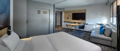 A hotel room with a large bed, a desk with a chair, a sofa with cushions, a flat-screen TV, and a window.