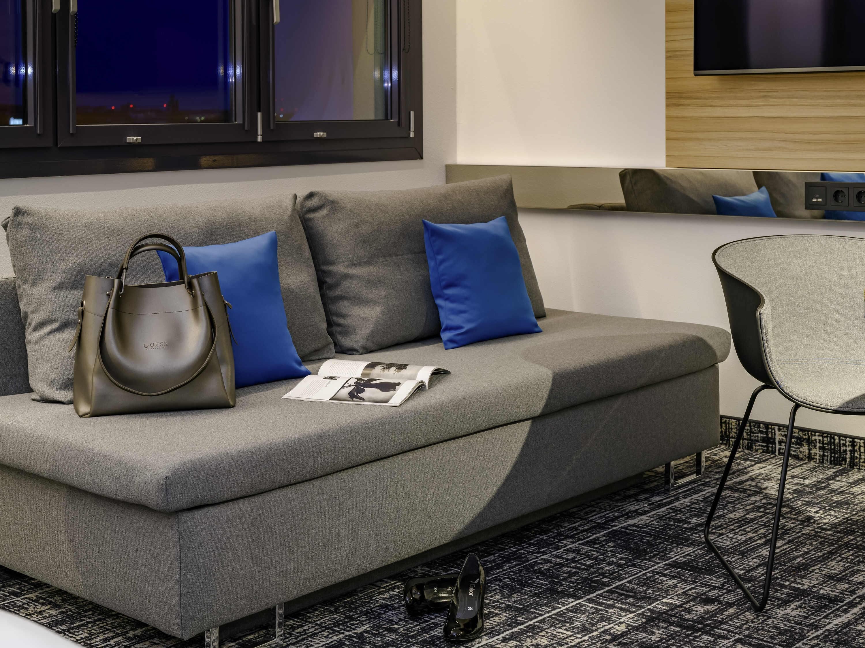 Une chambre d’hôtel moderne avec un canapé gris, des coussins bleus, un fauteuil gris, un sac à main noir et un magazine posé sur le canapé.