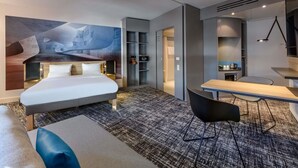 A modern hotel room with a large bed, a desk, a chair, and a shower area. - Novotel Suites Berlin City Potsdamer Platz (Berlin)