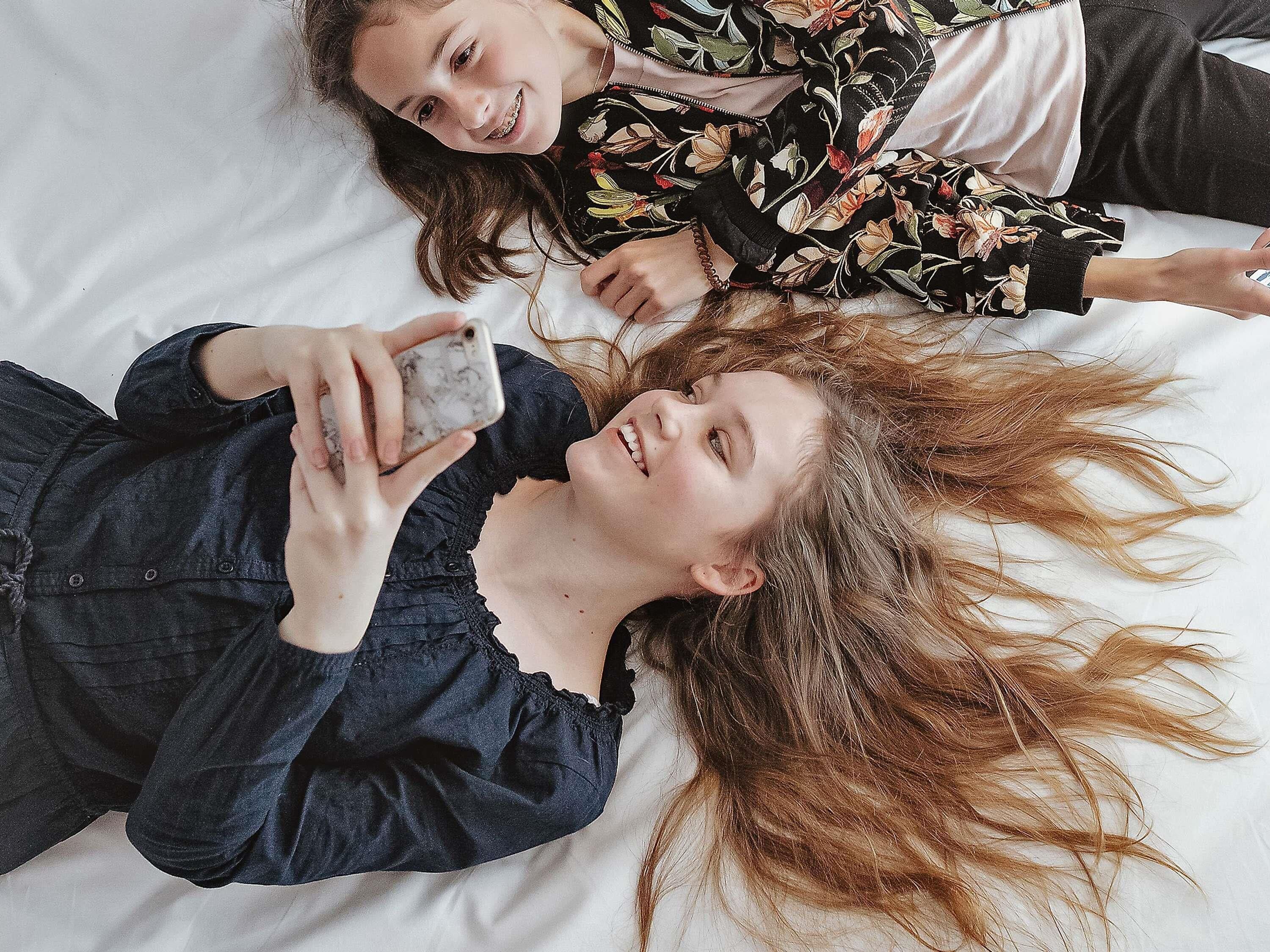 Dos personas recostadas en una cama con sábana blanca, una sosteniendo un teléfono inteligente.