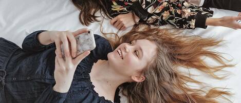 Dos personas recostadas en una cama con sábana blanca, una sosteniendo un teléfono inteligente.