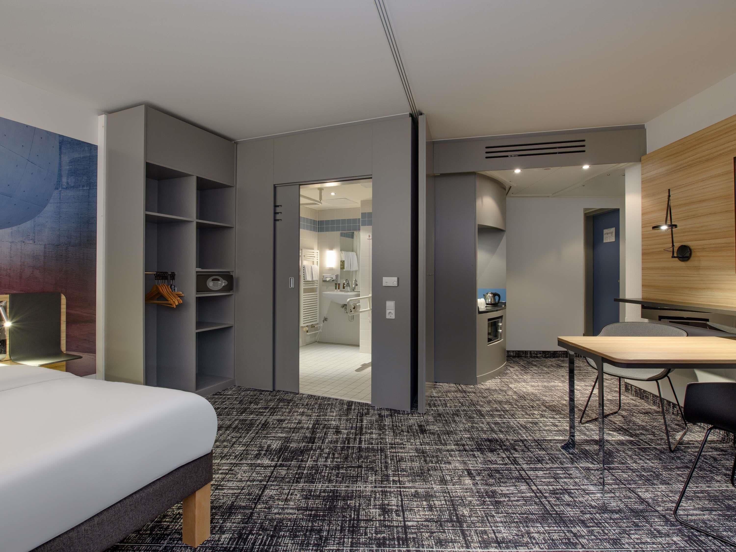 Une chambre d’hôtel moderne, équipée d’un lit, d’un bureau, d’une chaise, d’une armoire et d’une salle de bain visible à travers une porte ouverte.