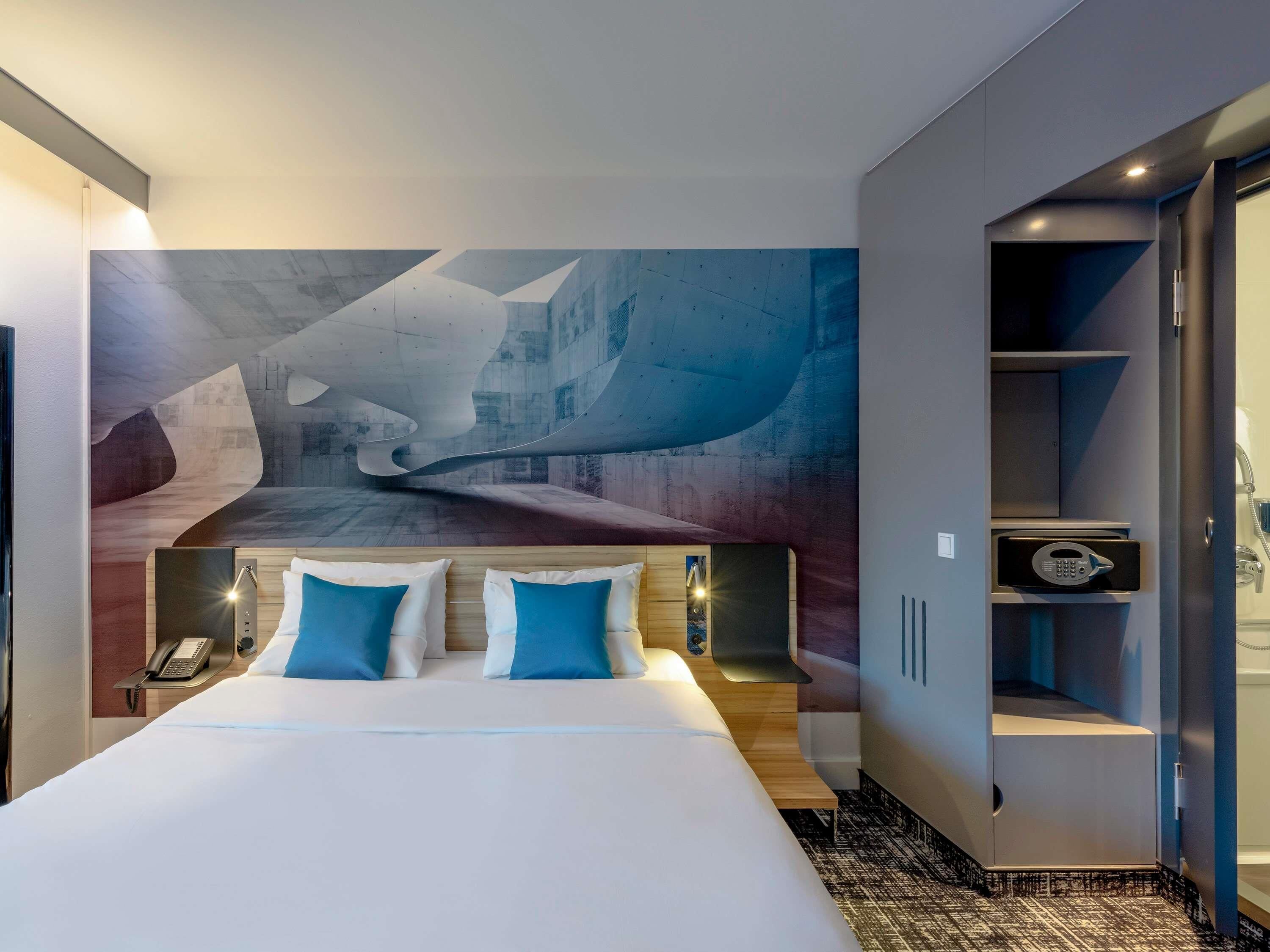 A modern hotel room with a large bed, a telephone, a safe, and a door leading to a bathroom.