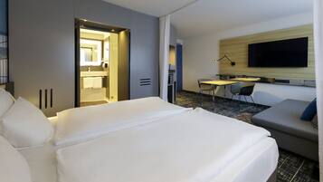 A hotel room with a large bed, a desk, a chair, a flat-screen TV, and a small seating area.