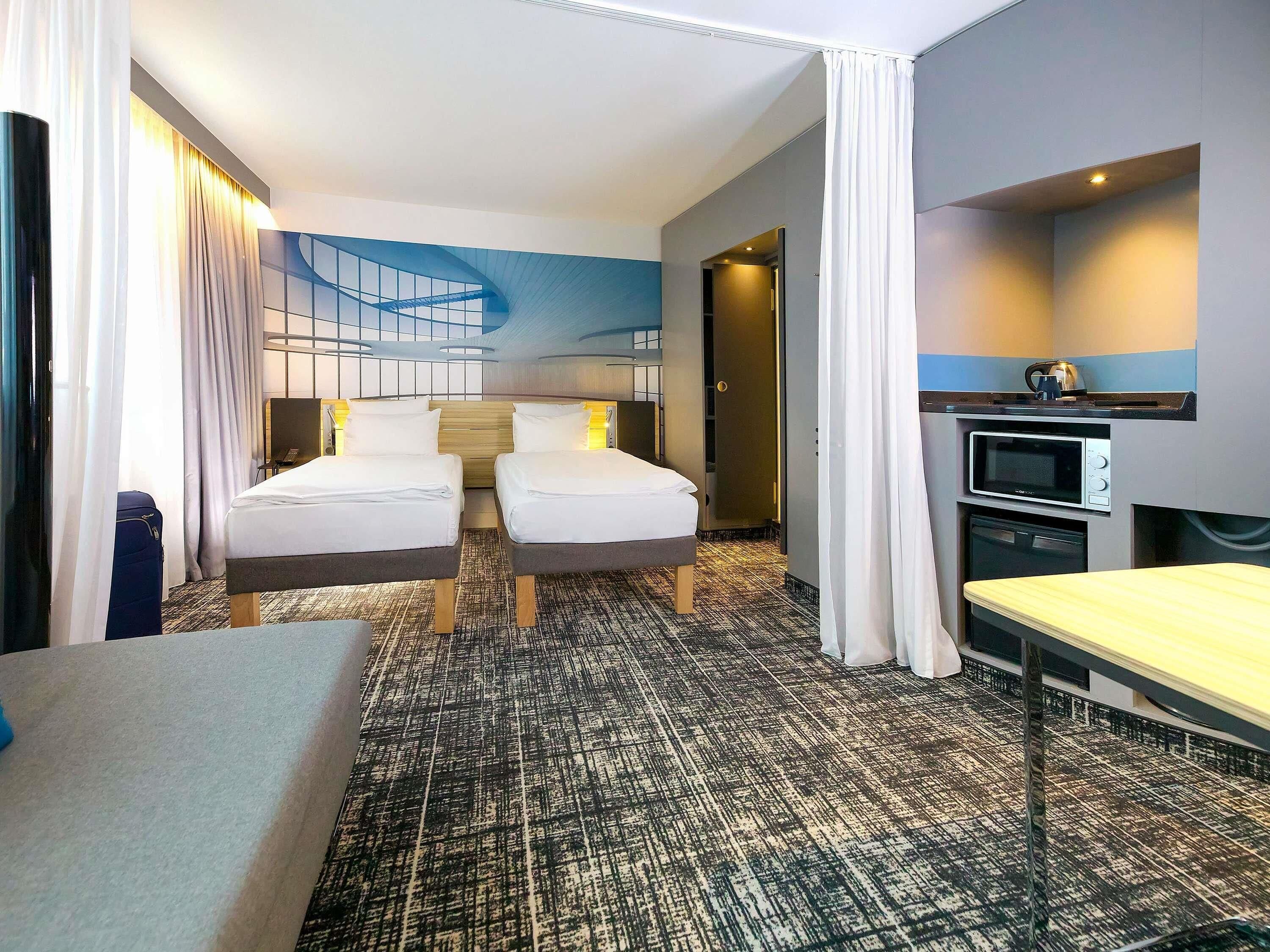 A hotel room with two beds, a small kitchenette, and a view of a building.