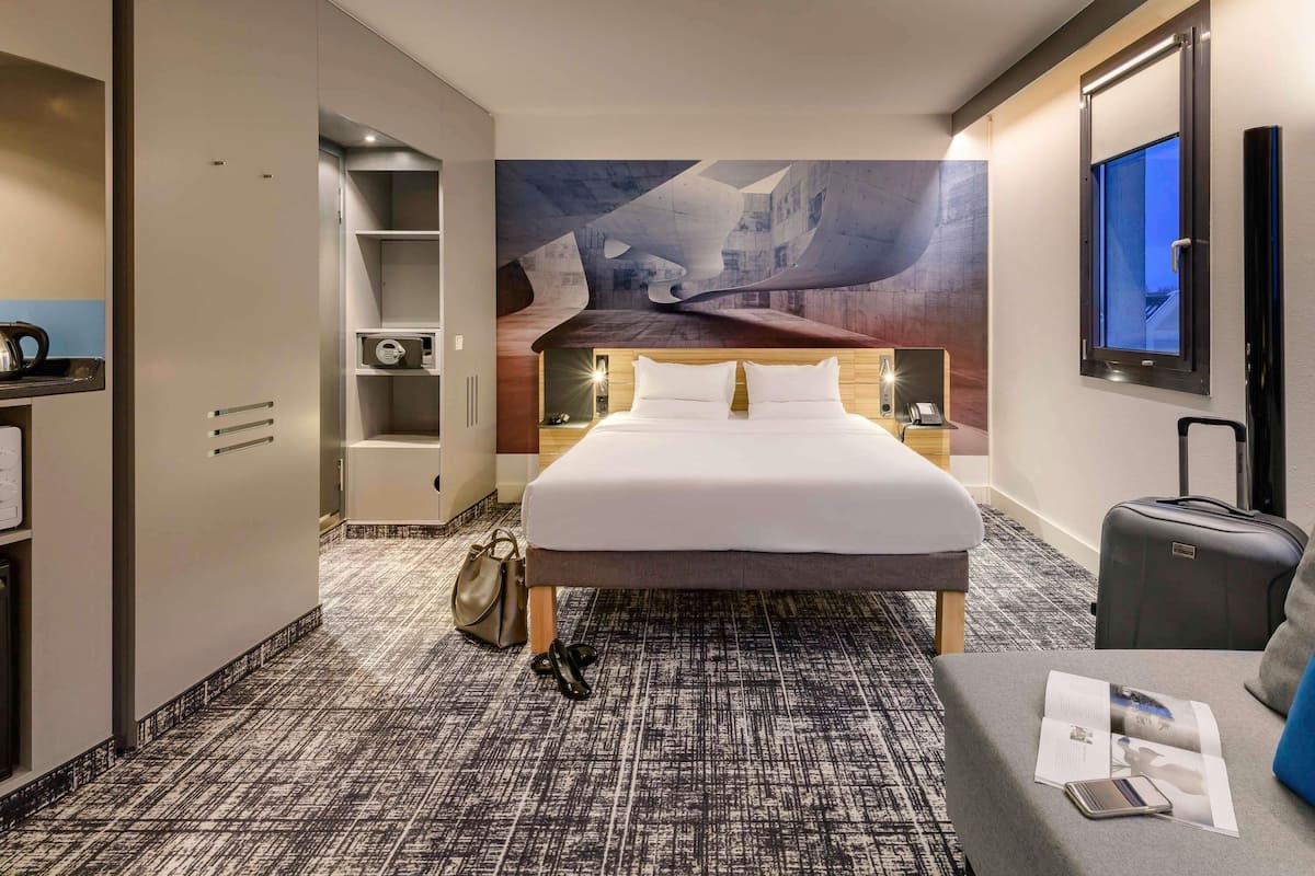 Habitación de hotel con cama, escritorio, armario, maleta y un cuadro en la pared.
