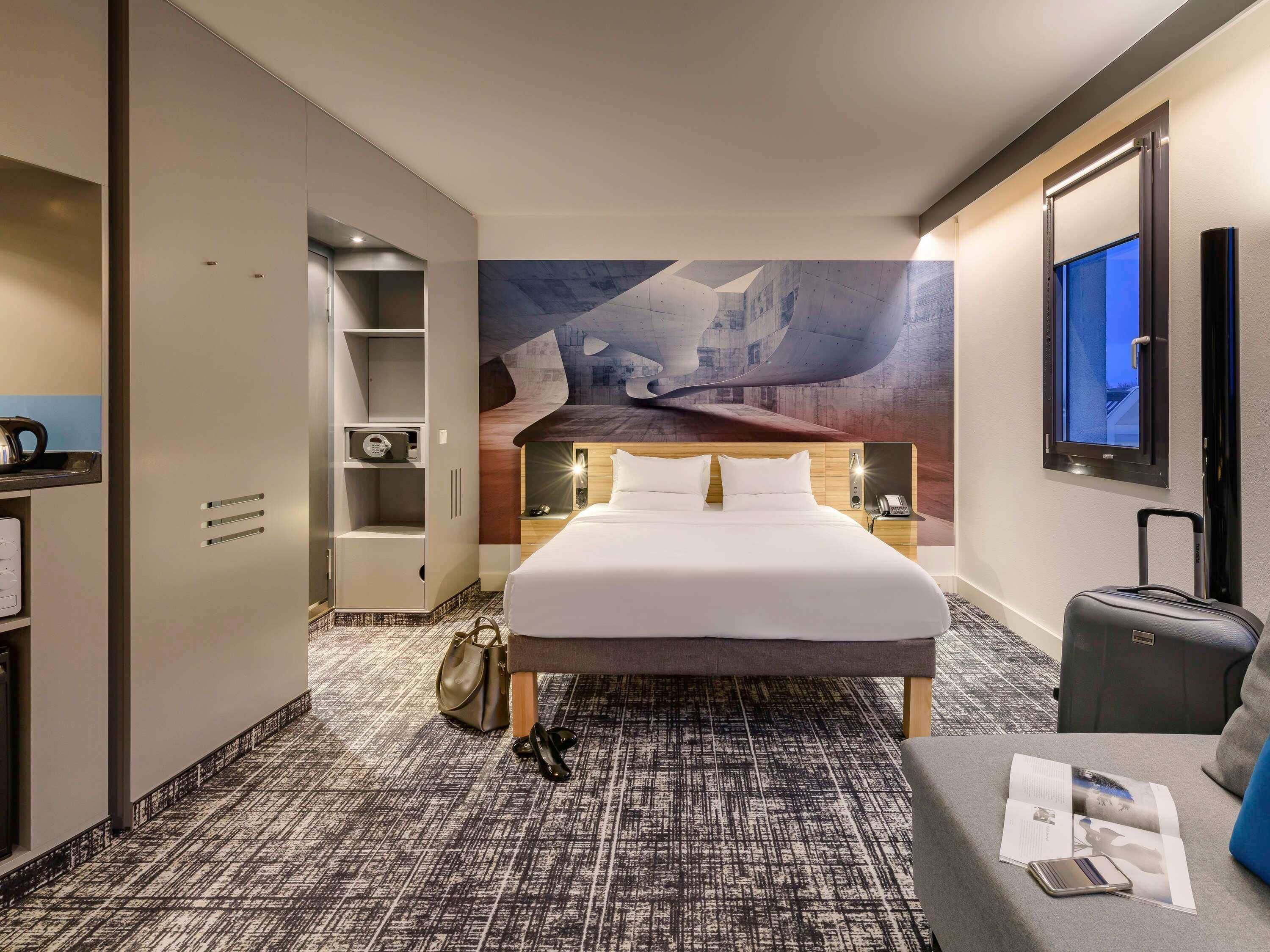 A hotel room with a bed, a desk, a wardrobe, a suitcase, and a wall with a painting.