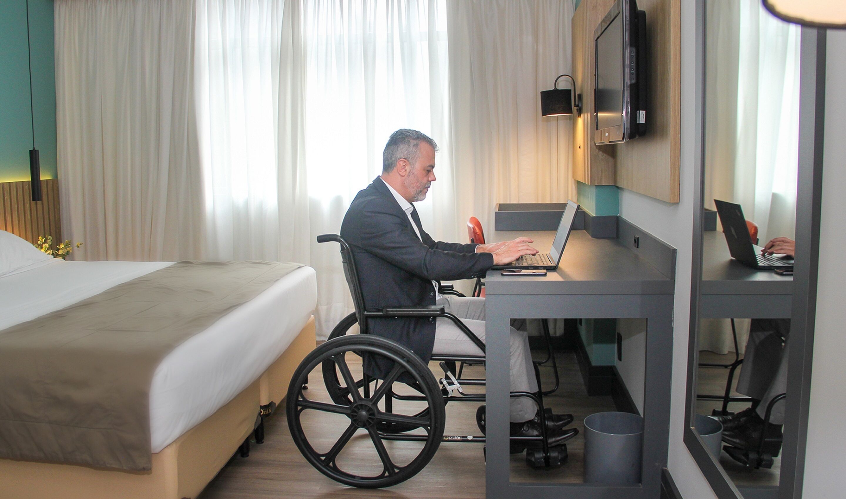 Ein Hotelzimmer mit einem Bett, einem Schreibtisch mit einem Laptop und einem Rollstuhl.