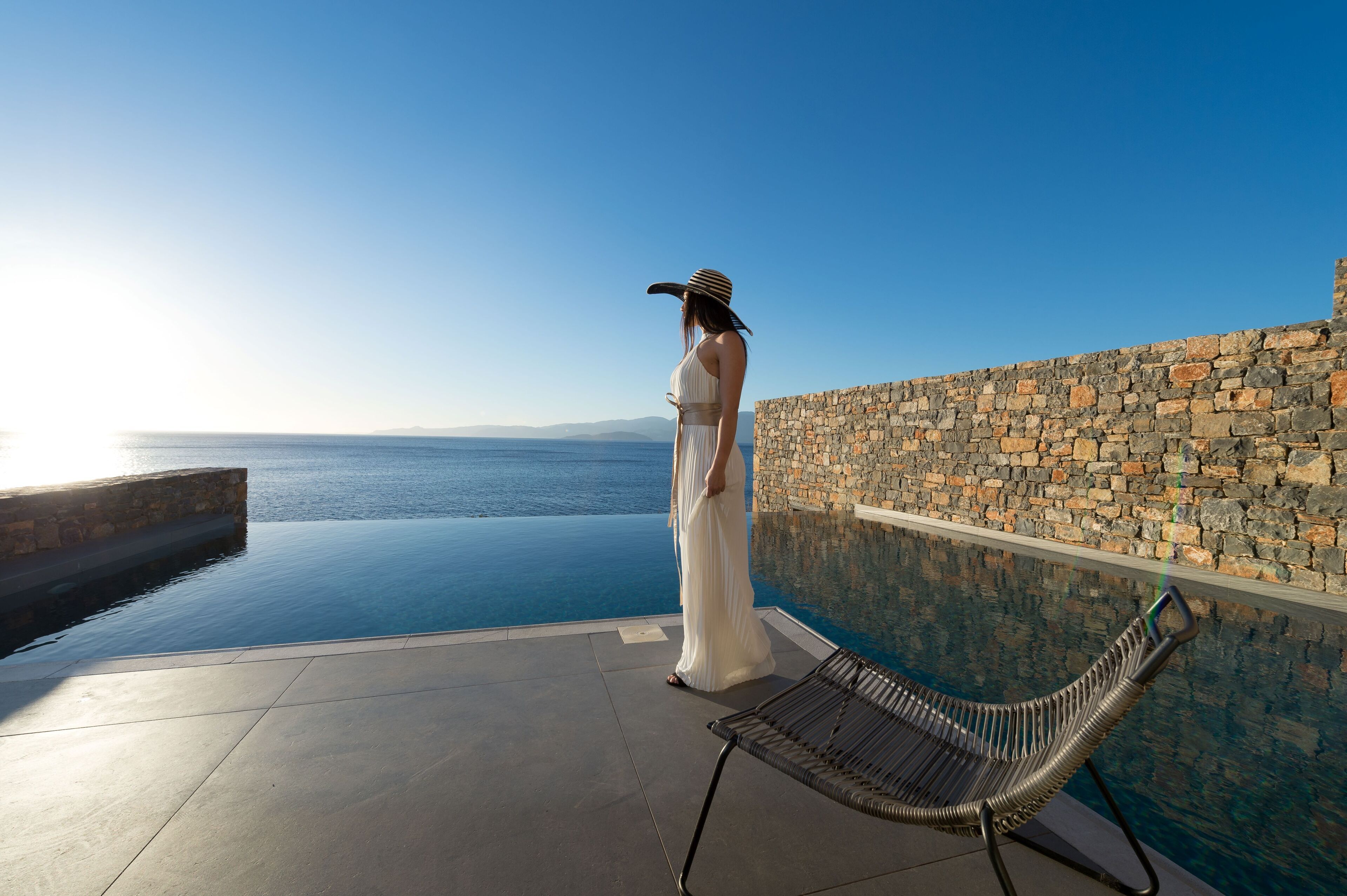 Una persona con vestido blanco de pie junto a una piscina infinita, con un muro de piedra y una silla metálica.