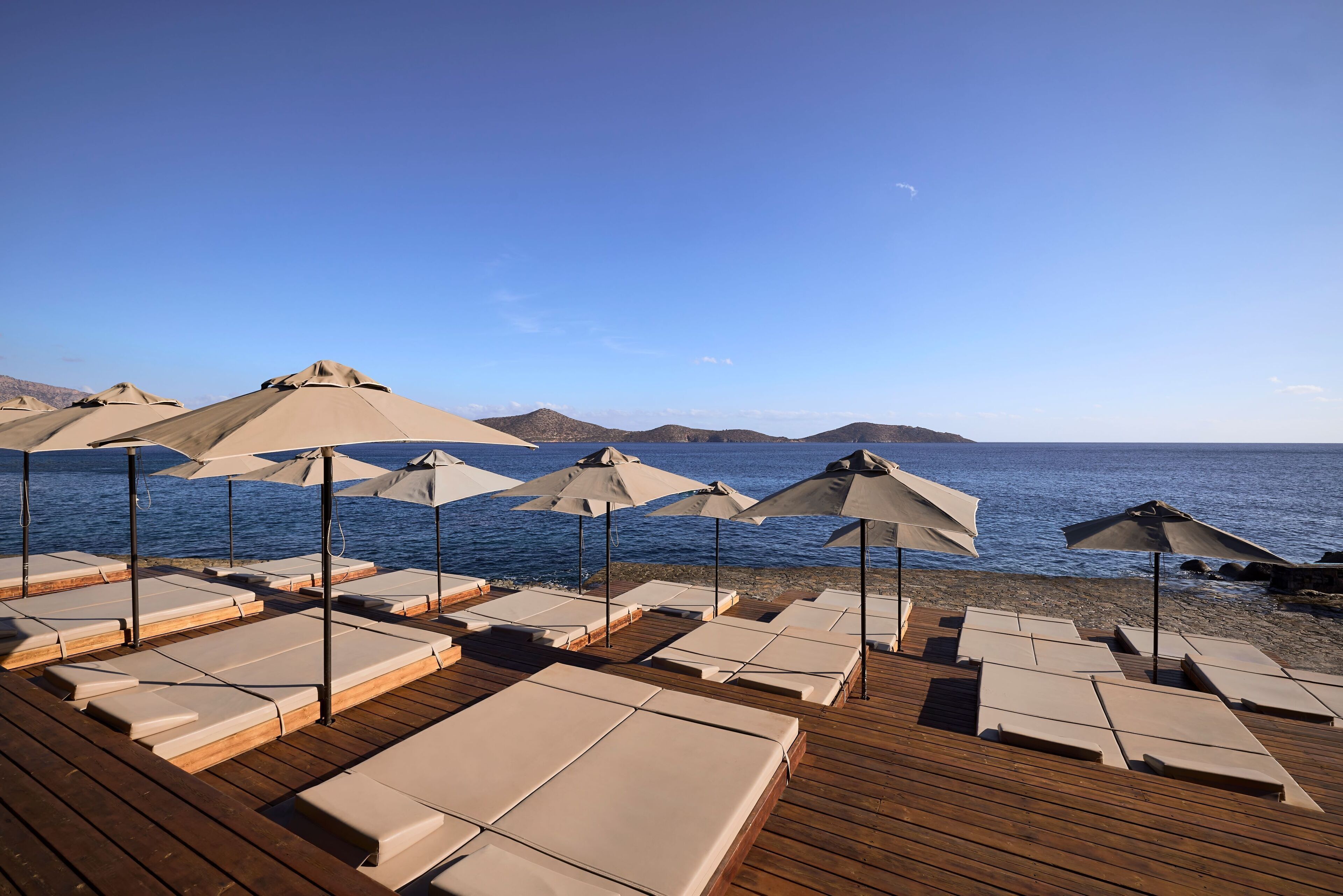 A beachside lounge area with sun loungers and umbrellas, wooden deck, and a view of the sea and distant hills.