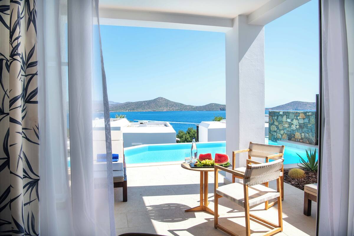 En balkon med udsigt over havet og bjergene, et bord med en vandmelon og en flaske, samt en stol.