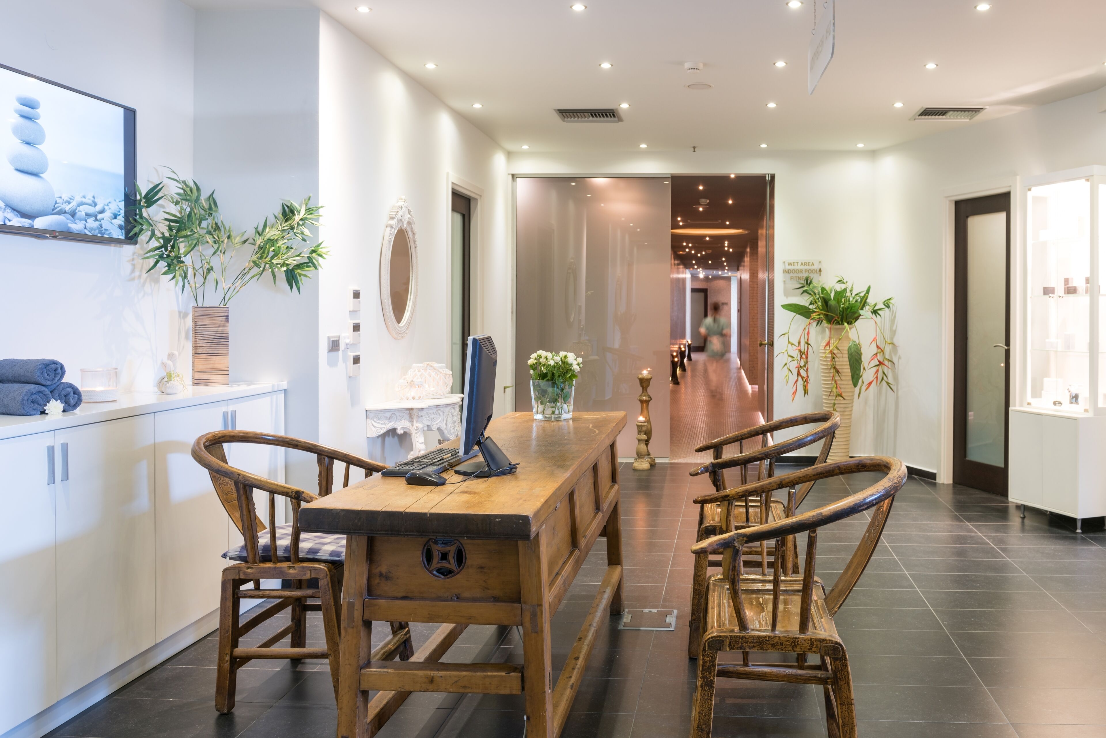 A modern hotel lobby with a wooden reception desk, two wooden chairs, a large mirror, and potted plants.