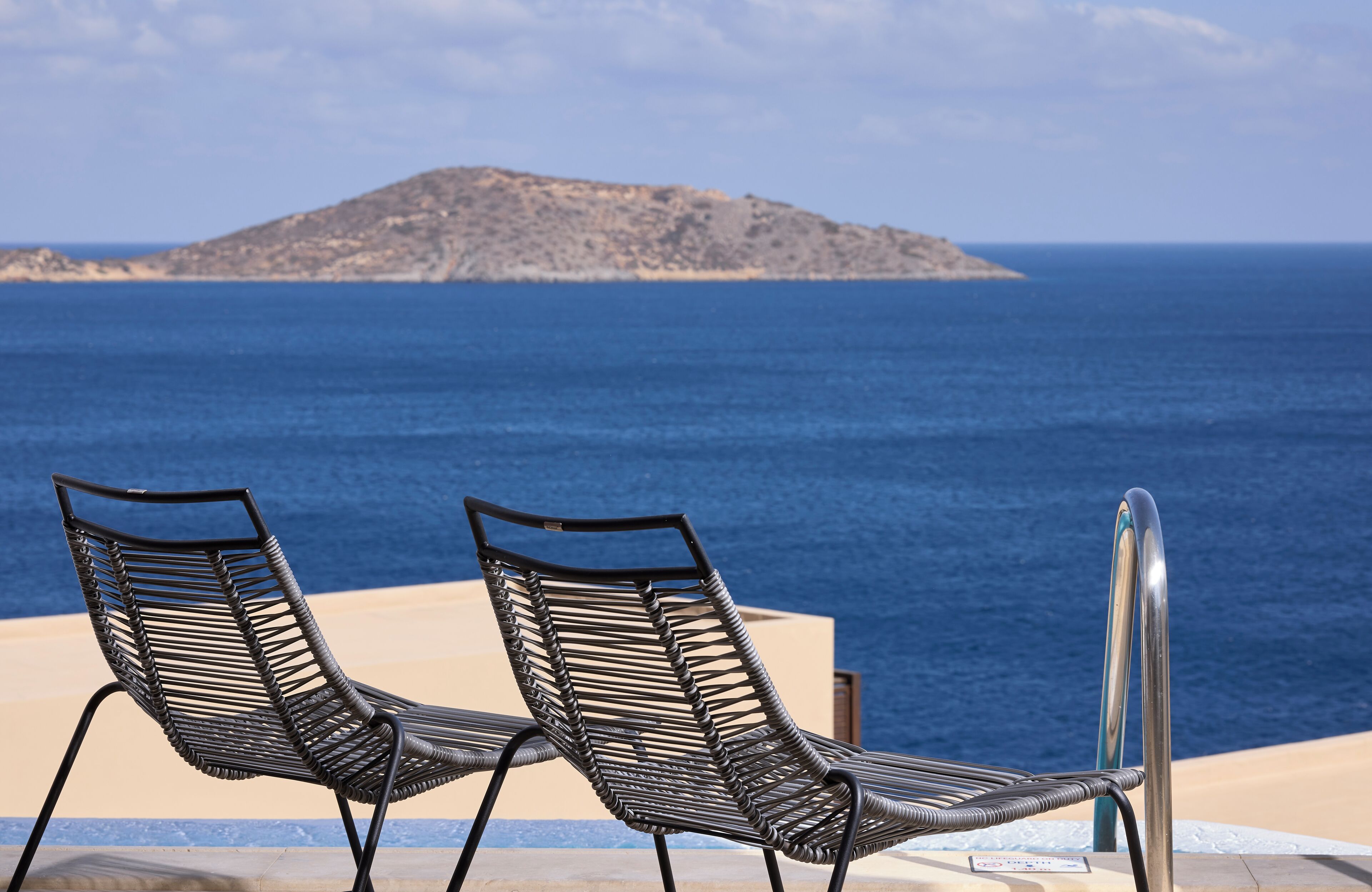 Two lounge chairs facing a pool with a view of the sea and a distant island.