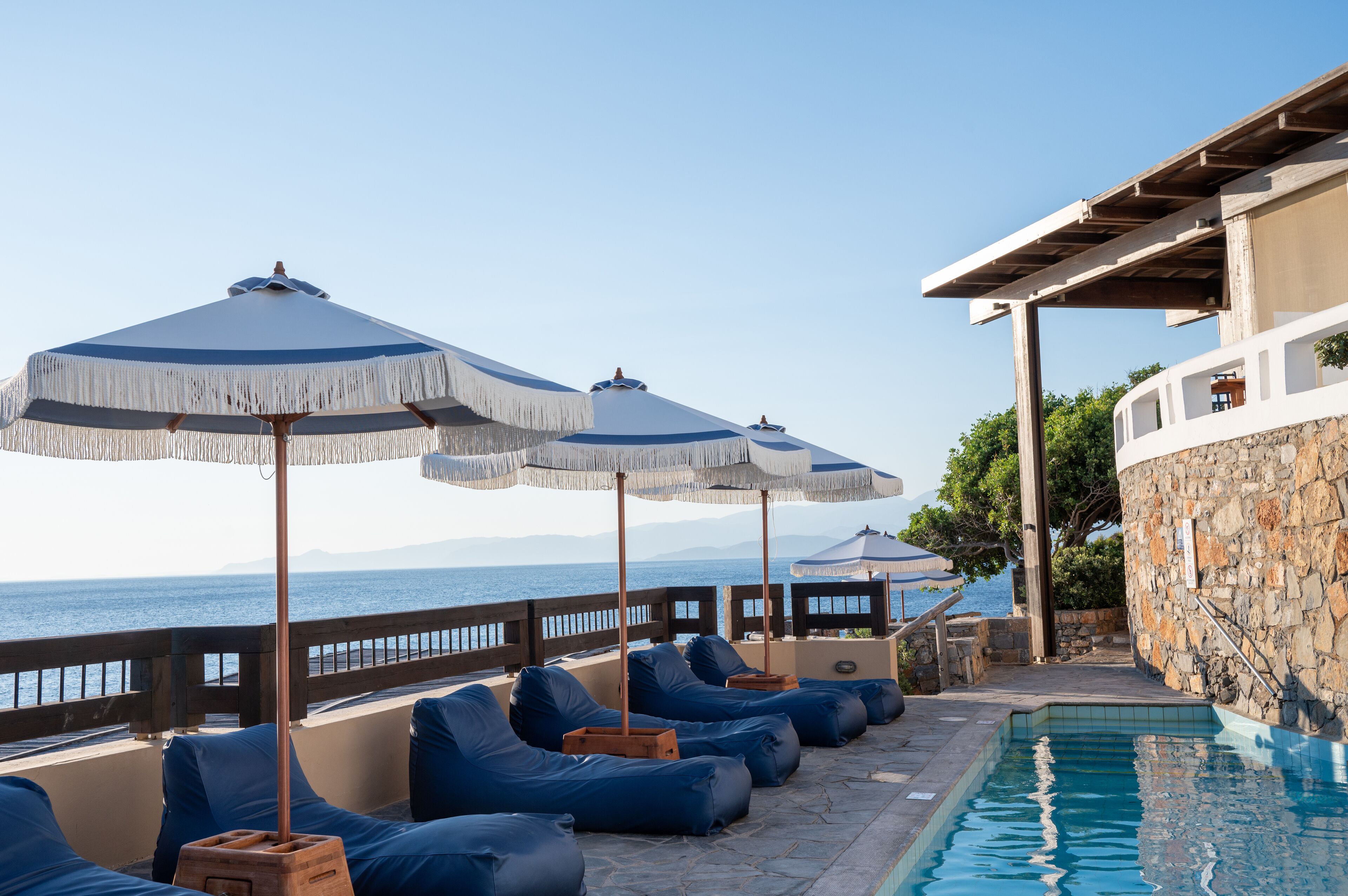 Zona de piscina con sillones de descanso y sombrillas, un muro de piedra y vista al mar.