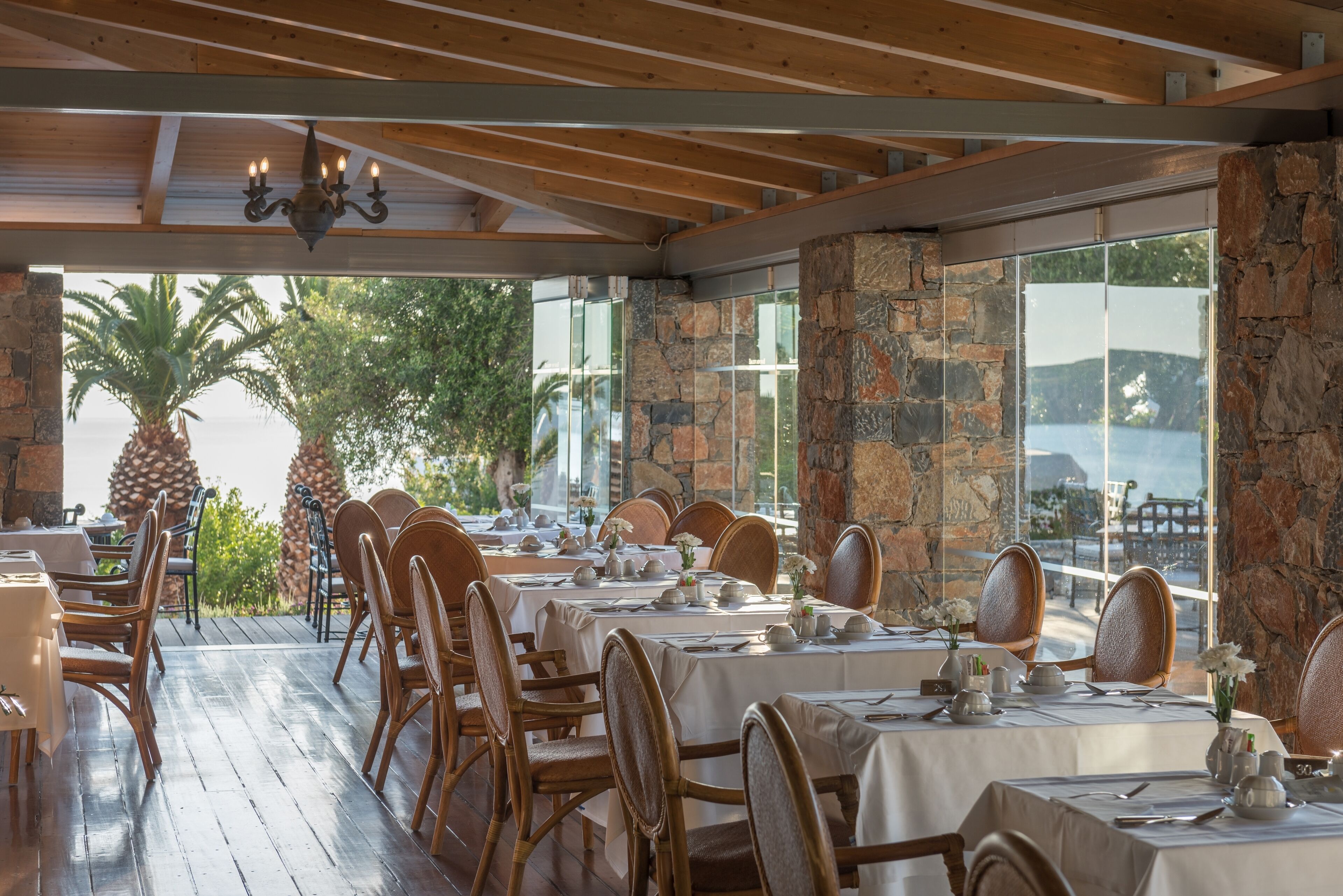 A restaurant with wooden tables and chairs, stone walls, and a view of the sea.