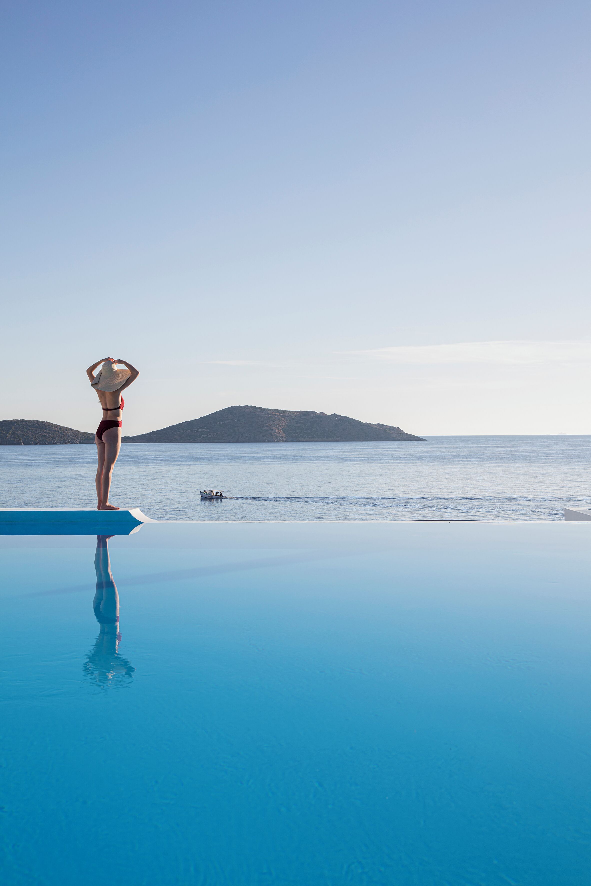 A person standing at the edge of a pool with a view of the sea and a boat in the distance.