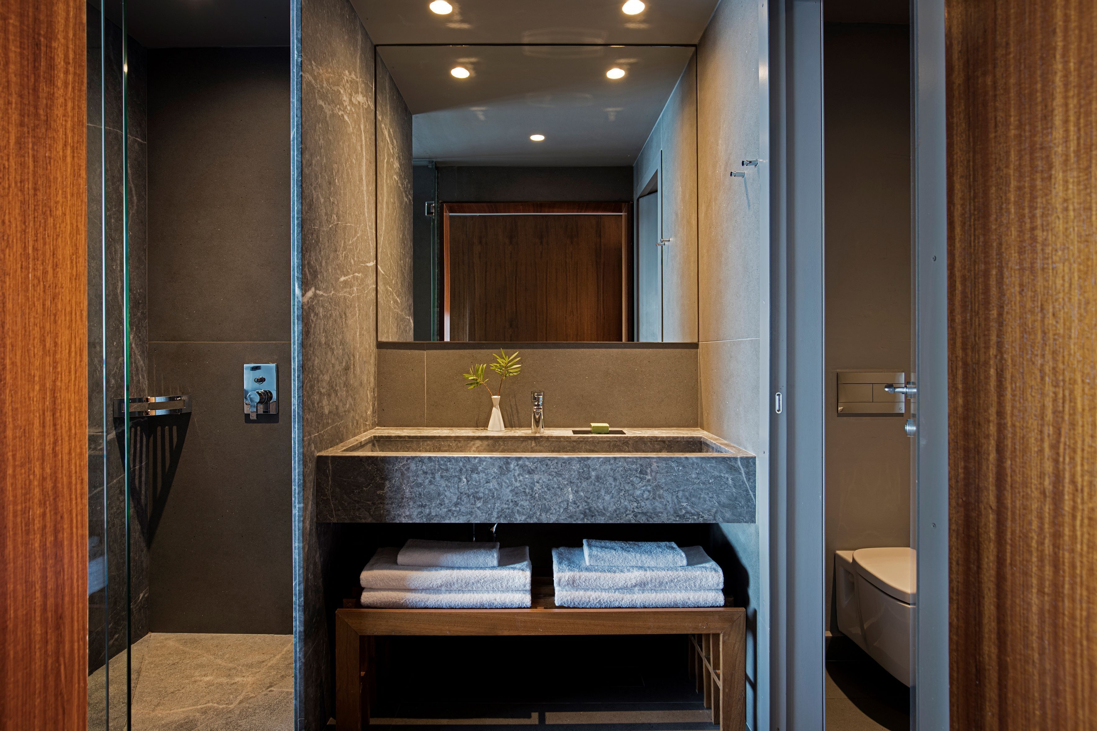Un baño moderno con lavamanos de mármol, banco de madera y un espejo grande.