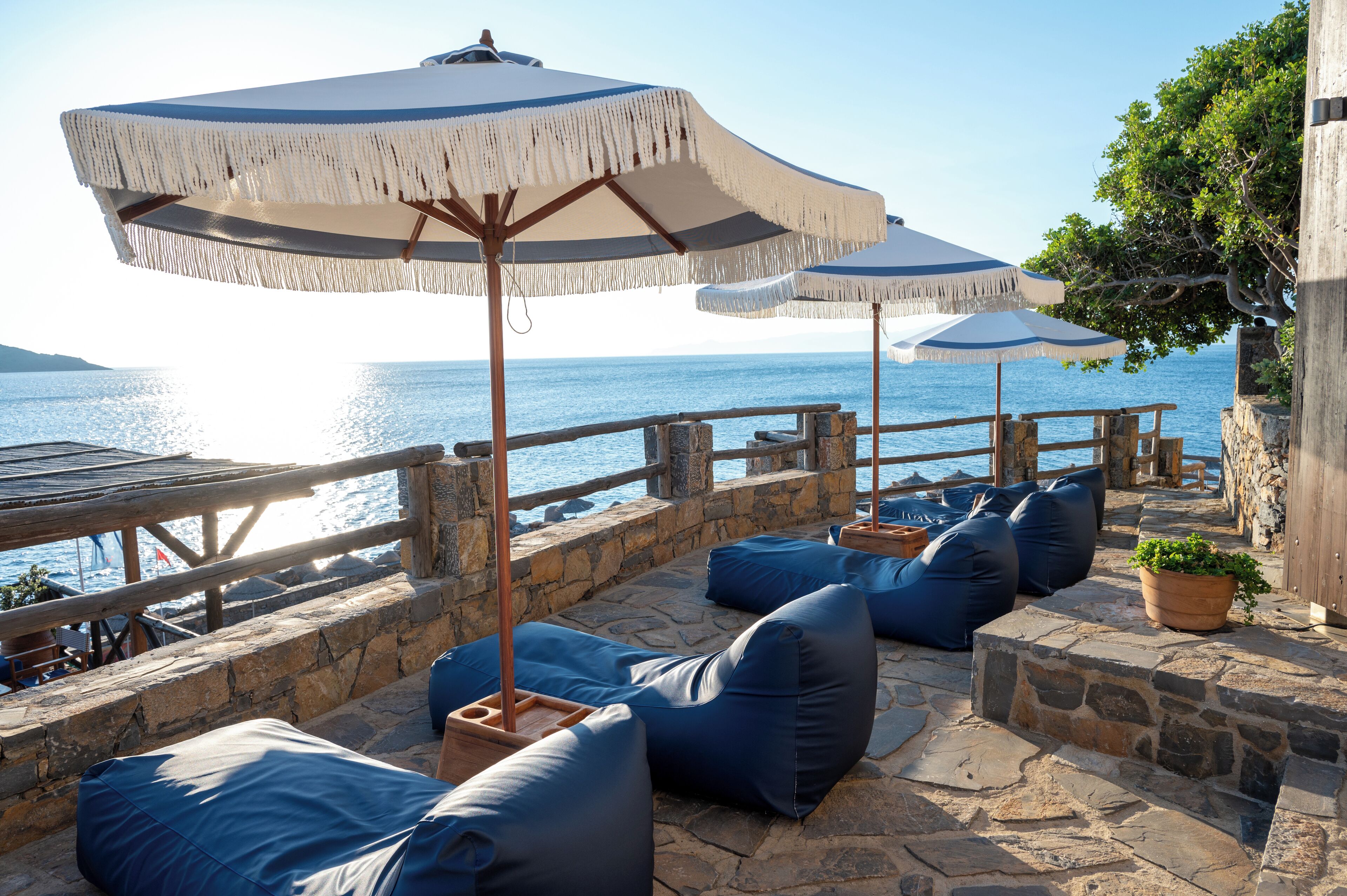 A seaside patio with stone flooring, blue cushioned seating, and large umbrellas.