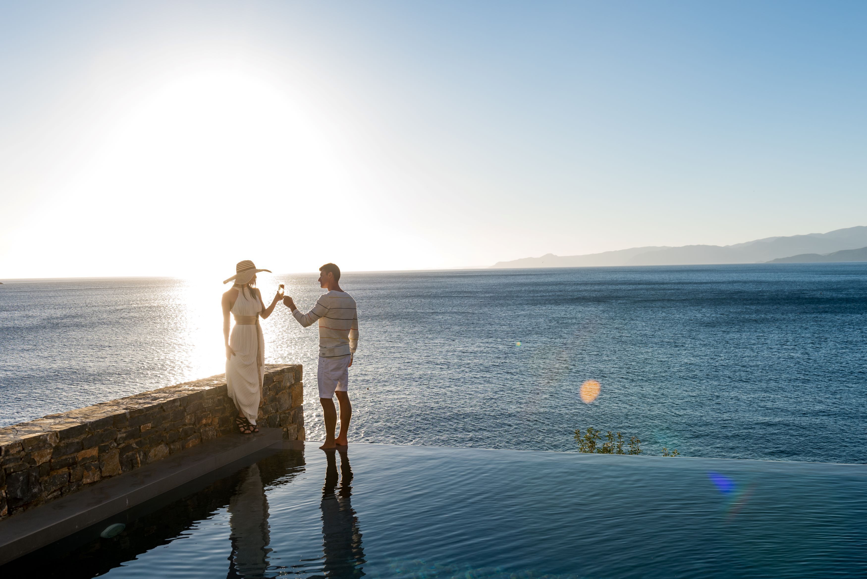 A couple standing by an infinity pool overlooking a body of water with a clear sky.