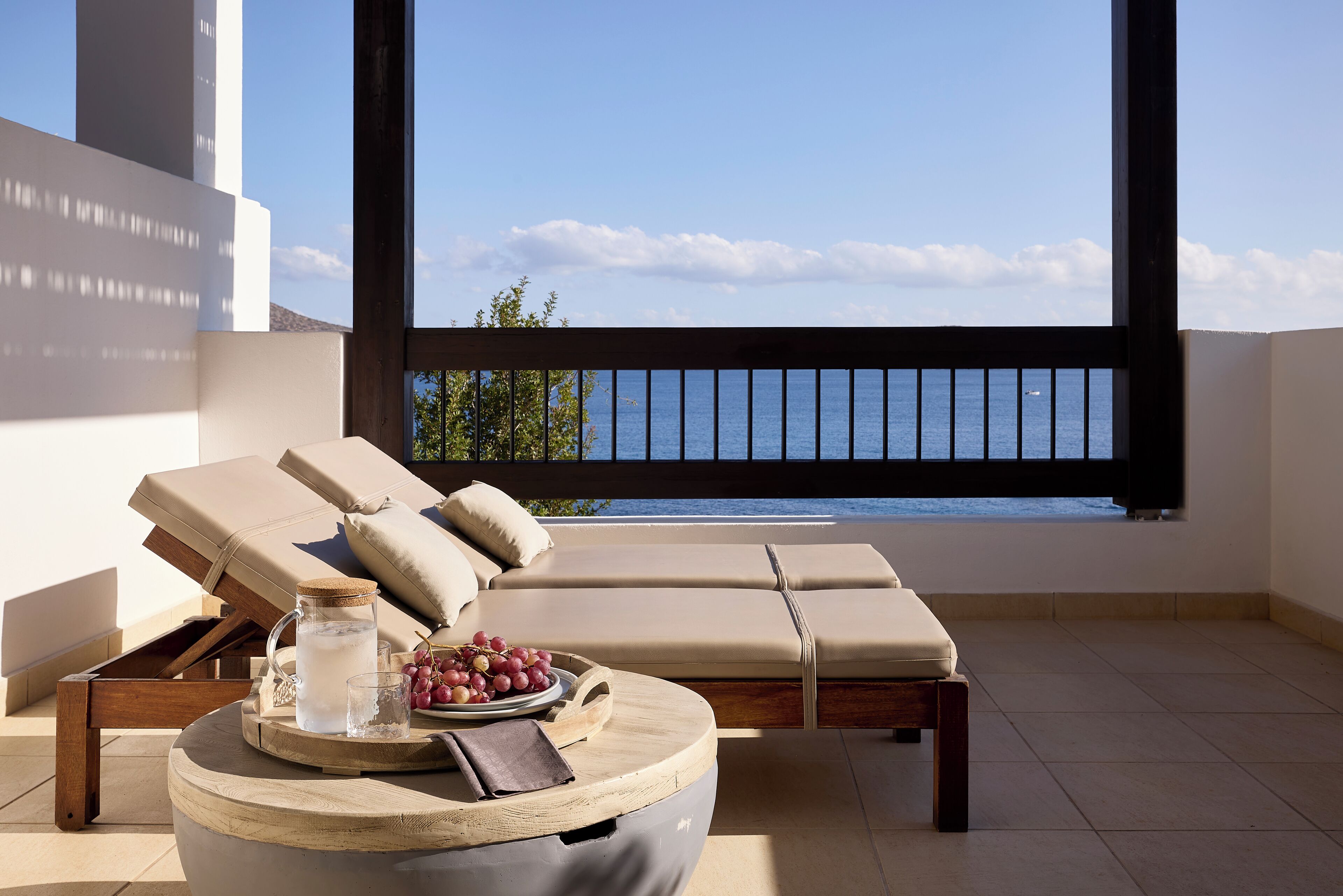 Ein sonnendurchfluteter Balkon mit Meerblick, eine Liege, ein kleiner Tisch mit einer Karaffe, Gläsern und Trauben.