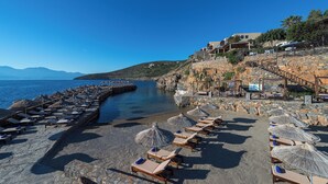Un hôtel en bord de mer, doté d’une architecture en pierre, d’une plage avec des transats et des parasols, et offrant une vue dégagée sur la mer.