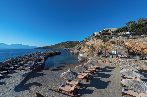 Un hôtel en bord de mer, doté d’une architecture en pierre, d’une plage avec des transats et des parasols, et offrant une vue dégagée sur la mer.