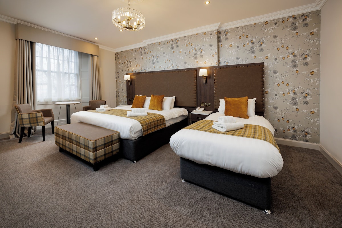 A hotel room with two beds, a chair, a table, and a chandelier.