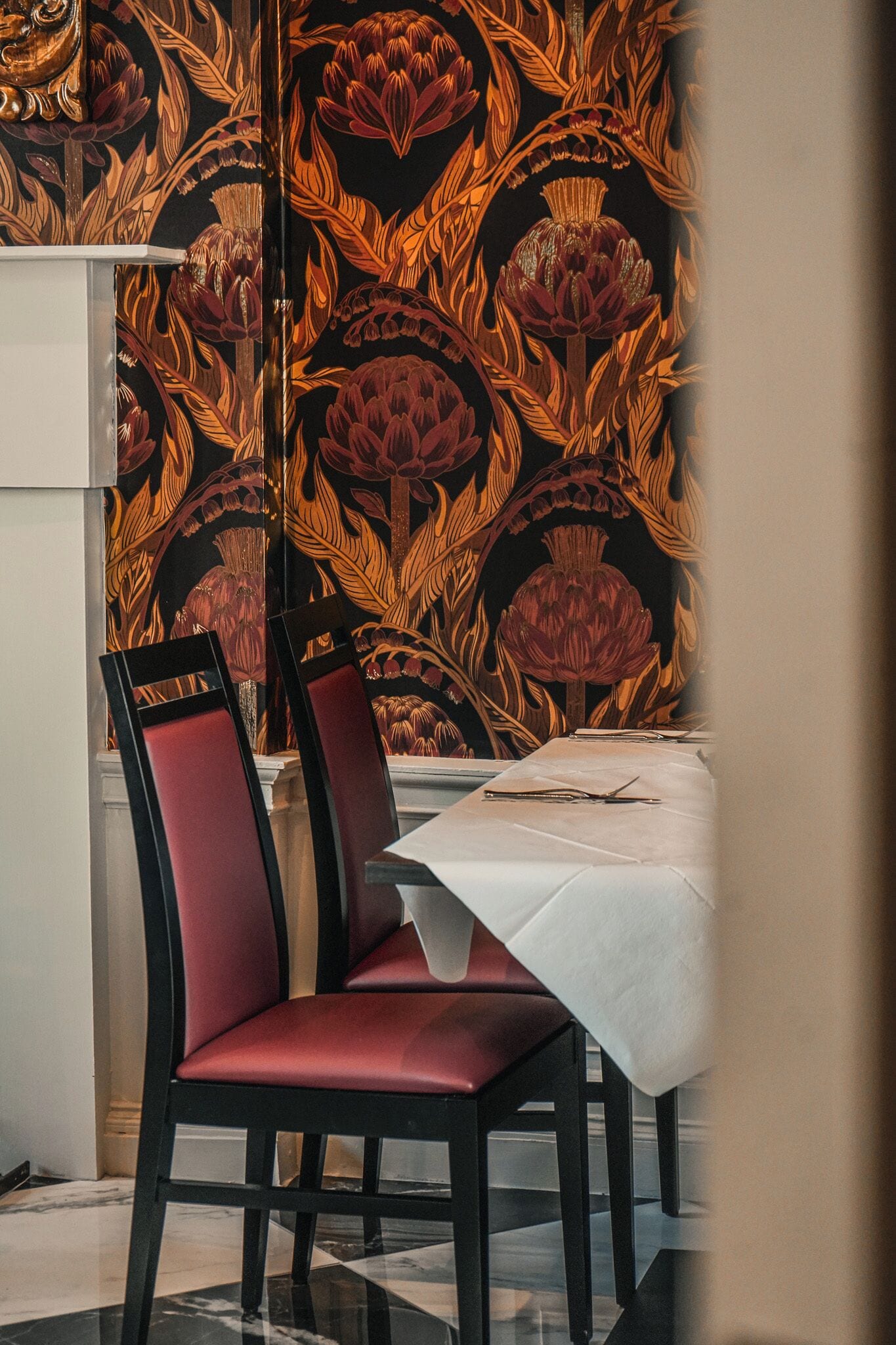 Un coin repas avec une table dressée pour deux personnes, une chaise avec un coussin rouge et un mur orné d’un motif floral.