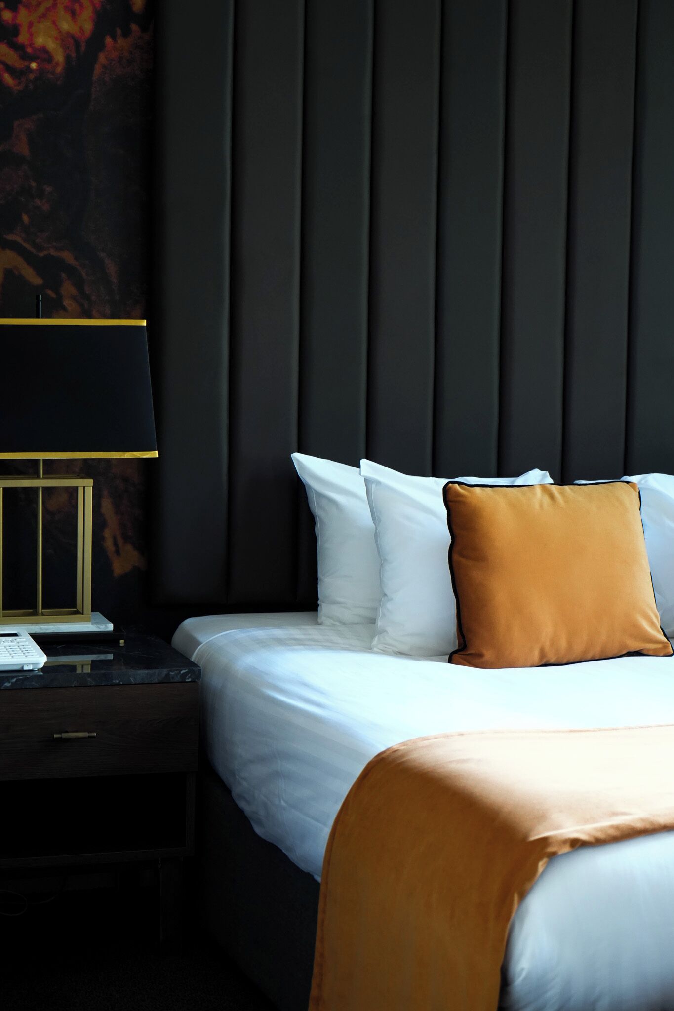 Een net opgemaakt bed met witte en bruine kussens, een donkere hoofdbord en een nachtkastje met een lamp.