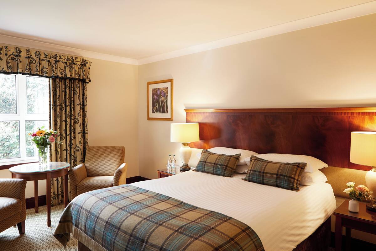 A hotel room with a large bed, two bedside lamps, a wooden headboard, a small table with a vase of flowers, a chair, and a window with curtains.