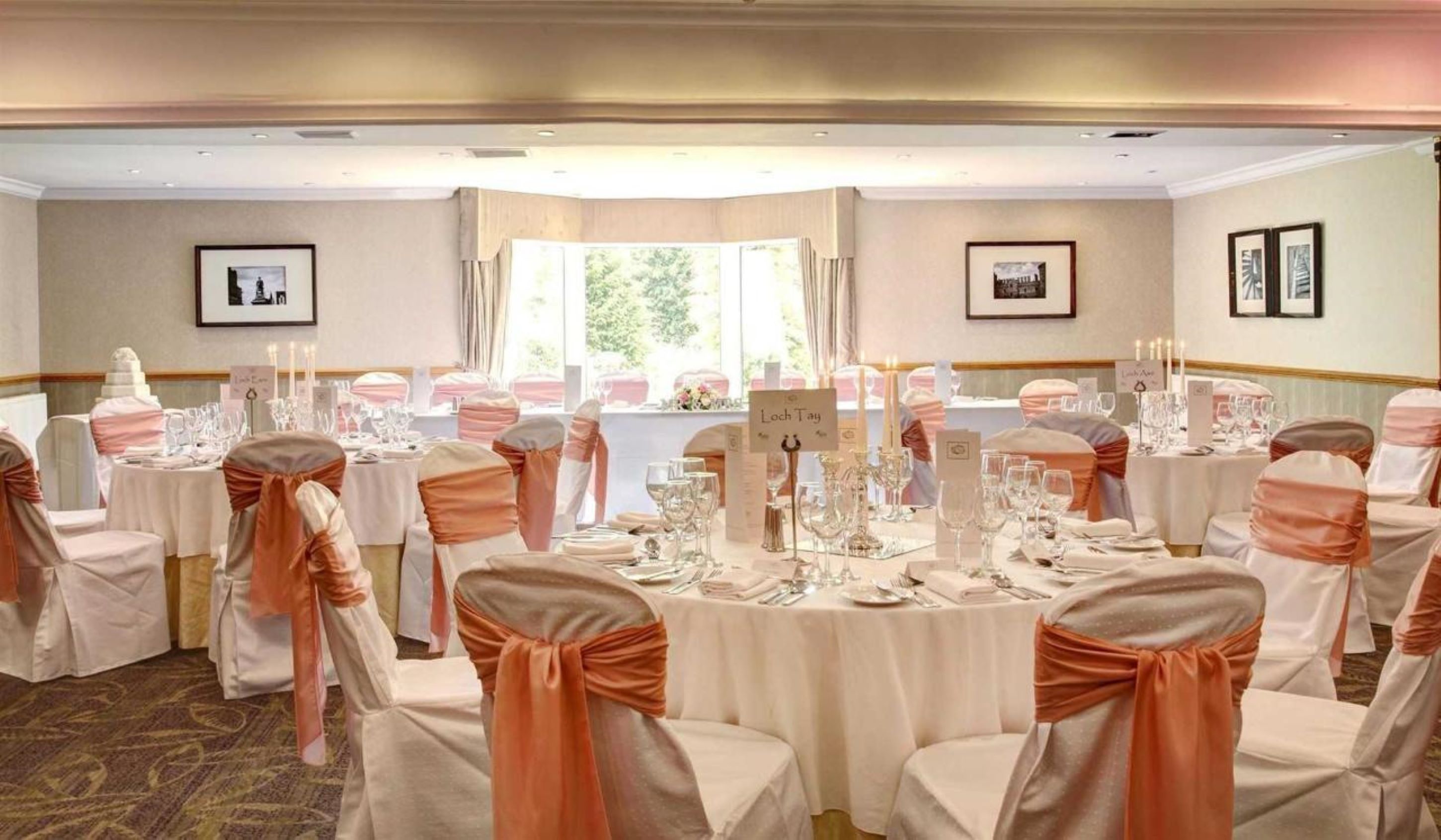 A banquet hall with round tables set for a formal event, featuring white tablecloths and orange sashes on chairs.