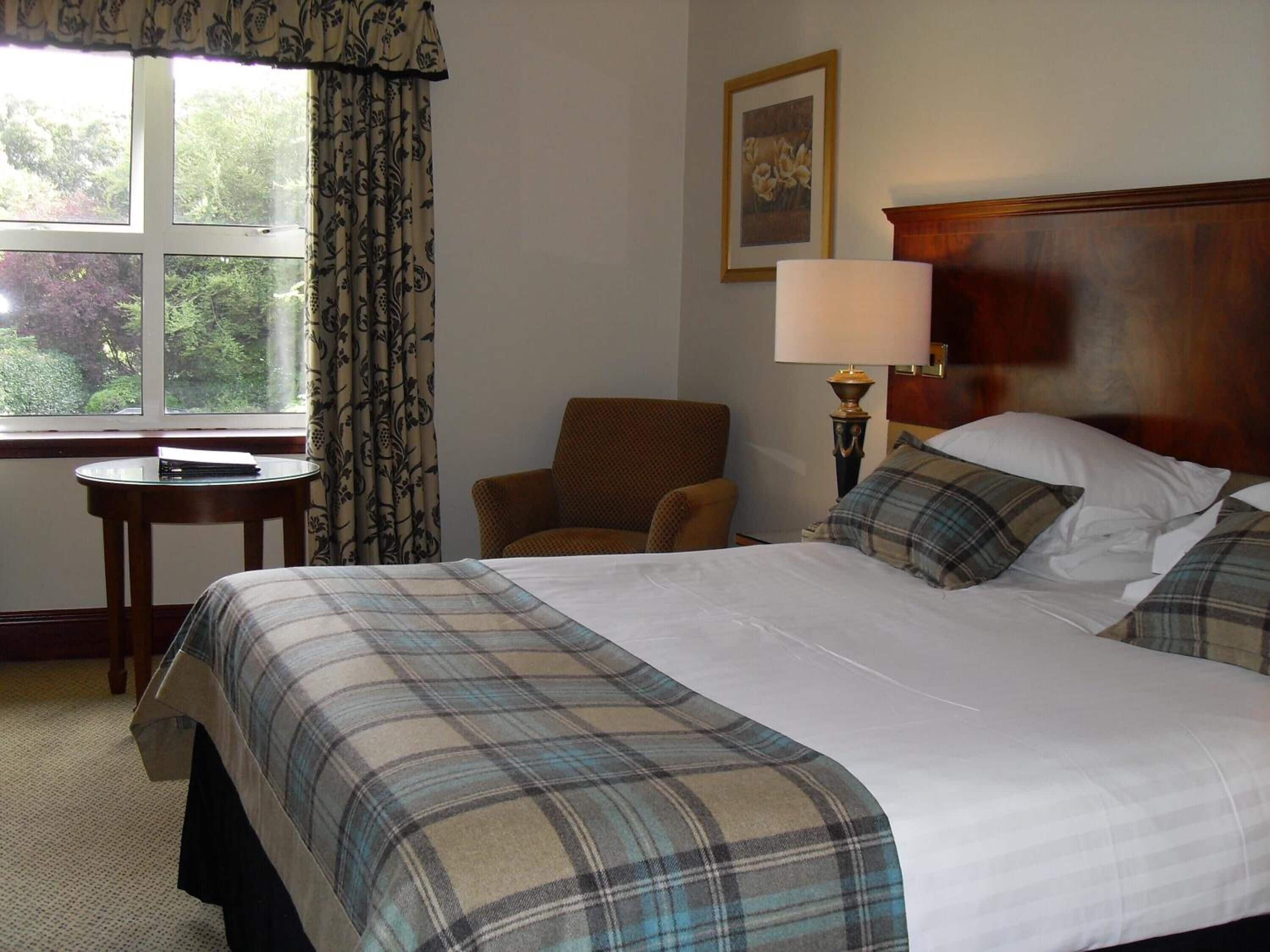Une chambre d’hôtel avec un grand lit, une chaise, une petite table, une lampe et une fenêtre avec des rideaux.