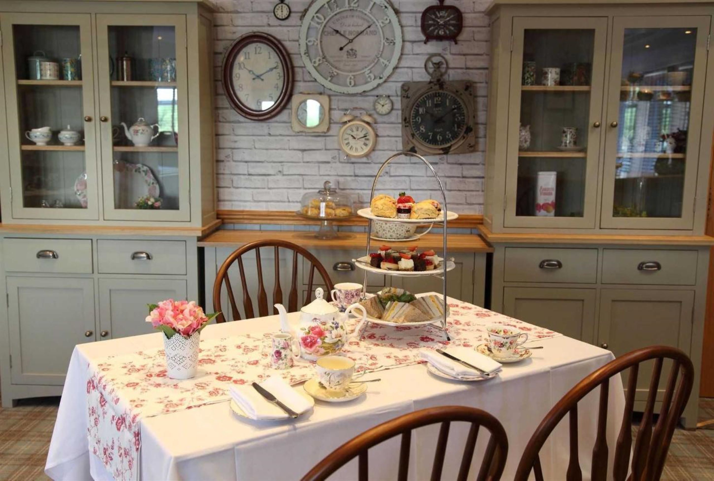 A dining area with a table set for four, surrounded by wooden chairs. There are two display cabinets with glass doors on either side of the table, and a wall clock above the cabinets.