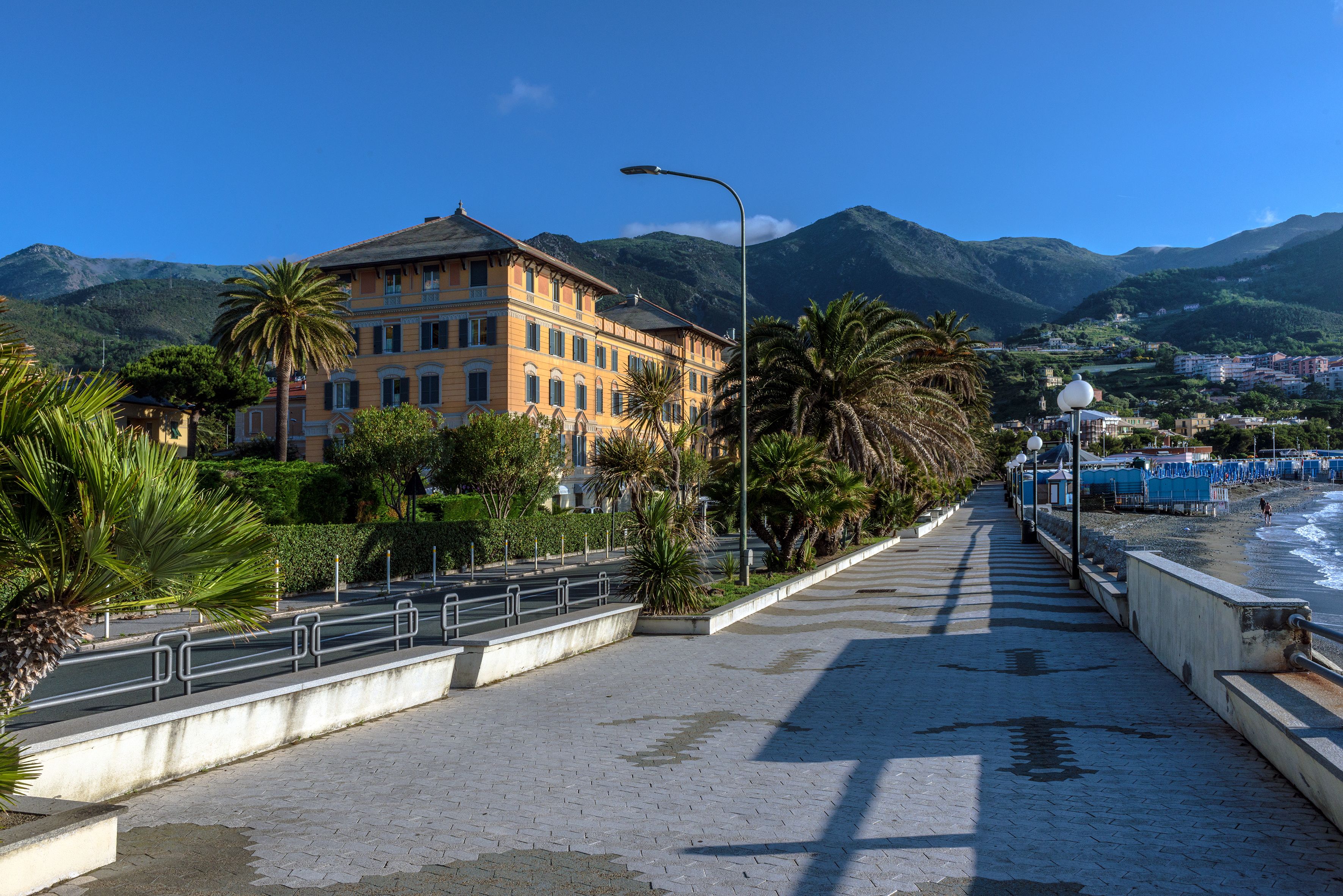 A coastal promenade with a building featuring balconies and a view of the sea and mountains.