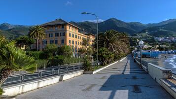 A coastal promenade with a building featuring balconies and a view of the sea and mountains.