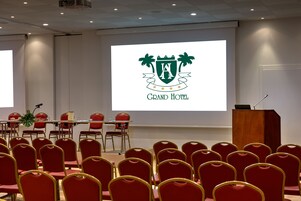 Konferenssihuone, jossa on punaiset tuolit, puhujapönttö ja näyttö, jossa näkyy hotellin logo.
