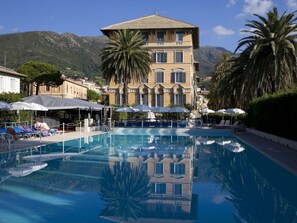 A pool area with lounge chairs and umbrellas, a building with a tiled roof, and palm trees. - Grand Hotel Arenzano (Arenzano)