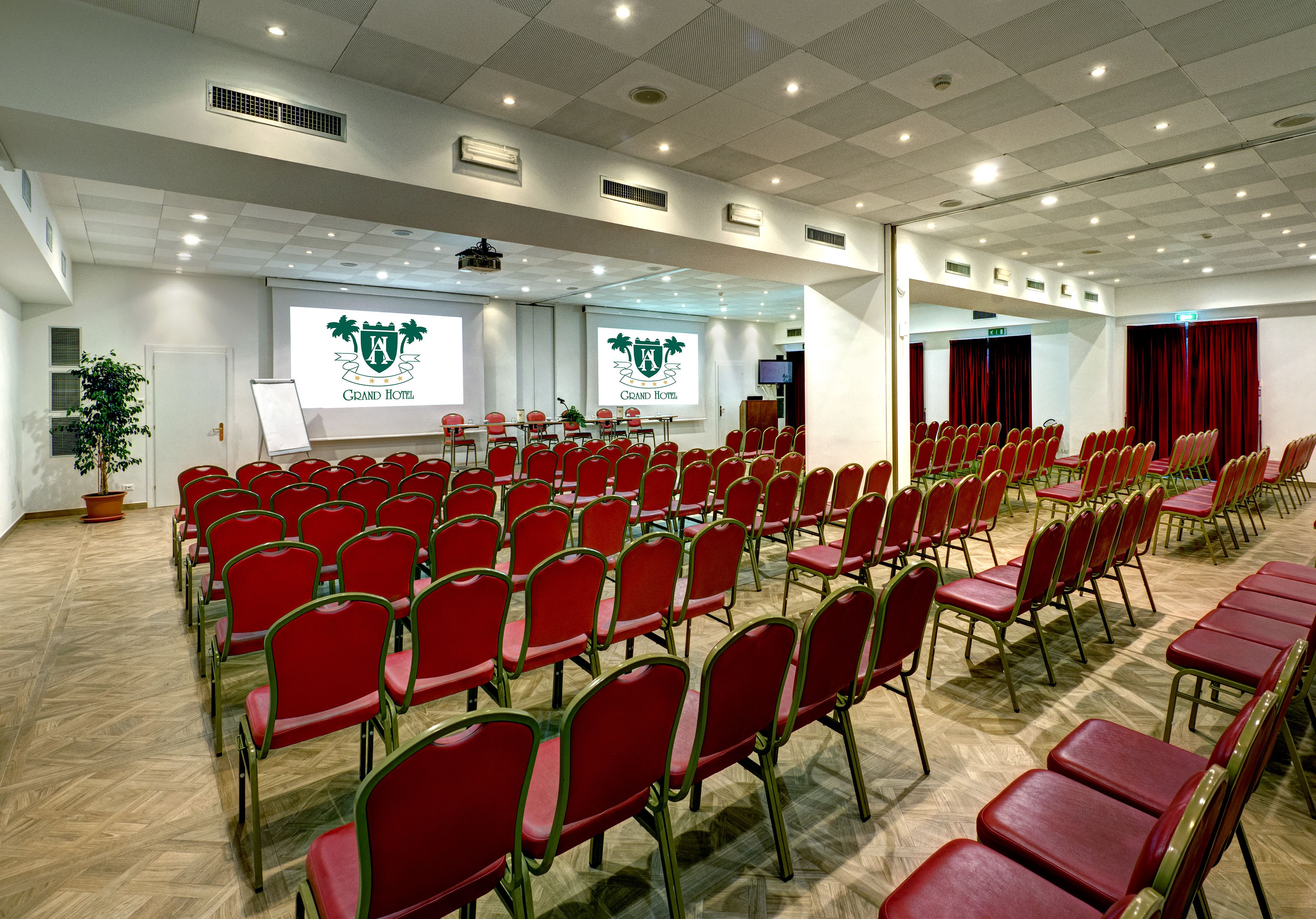 Une salle de conférence avec des rangées de chaises rouges, un écran de projection et un logo sur le mur.