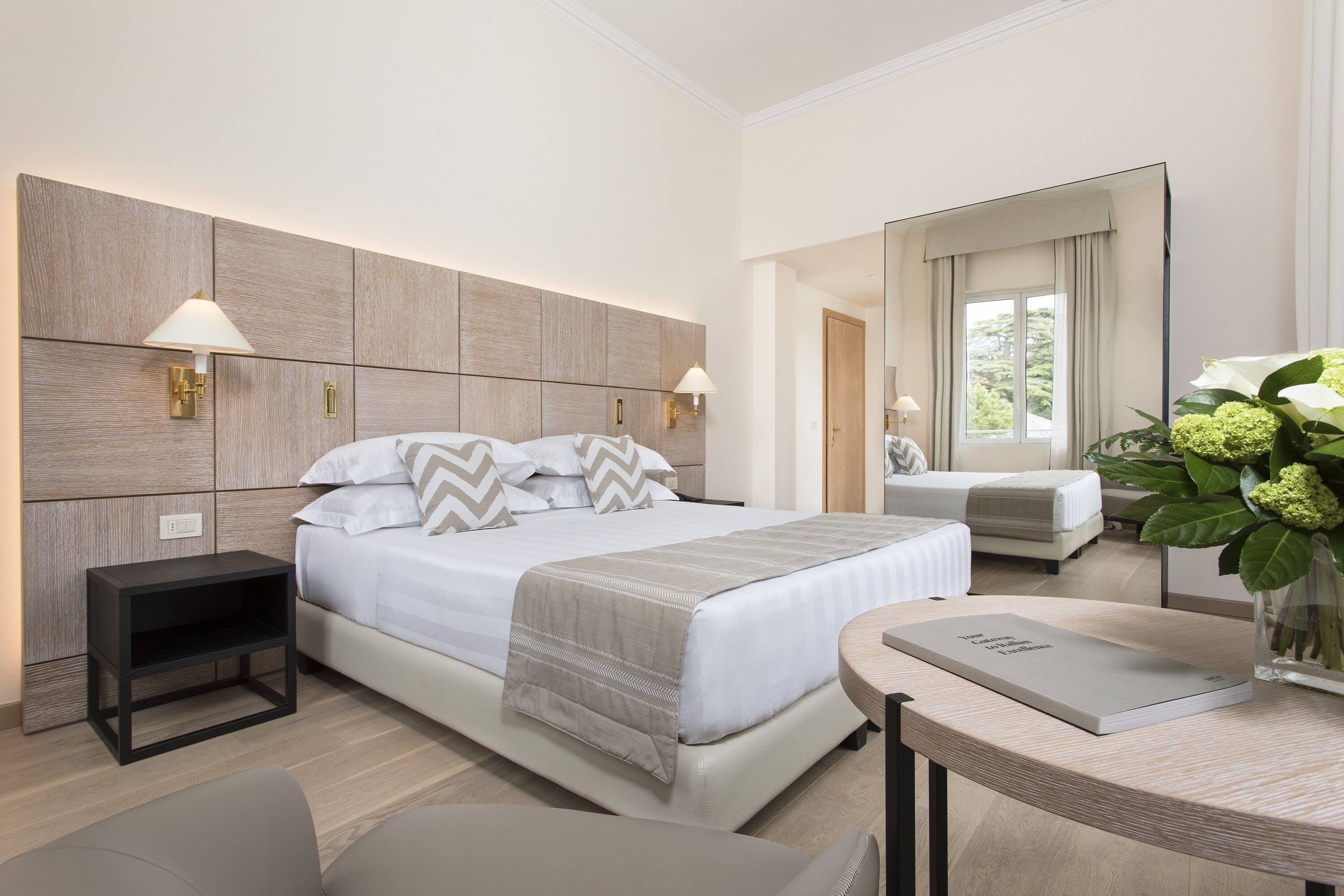 Ein modernes Hotelzimmer mit einem großen Bett, Nachttischlampen, einem Nachttisch, einem kleinen Beistelltisch, einem runden Tisch mit einem Buch und einer Vase mit Blumen.