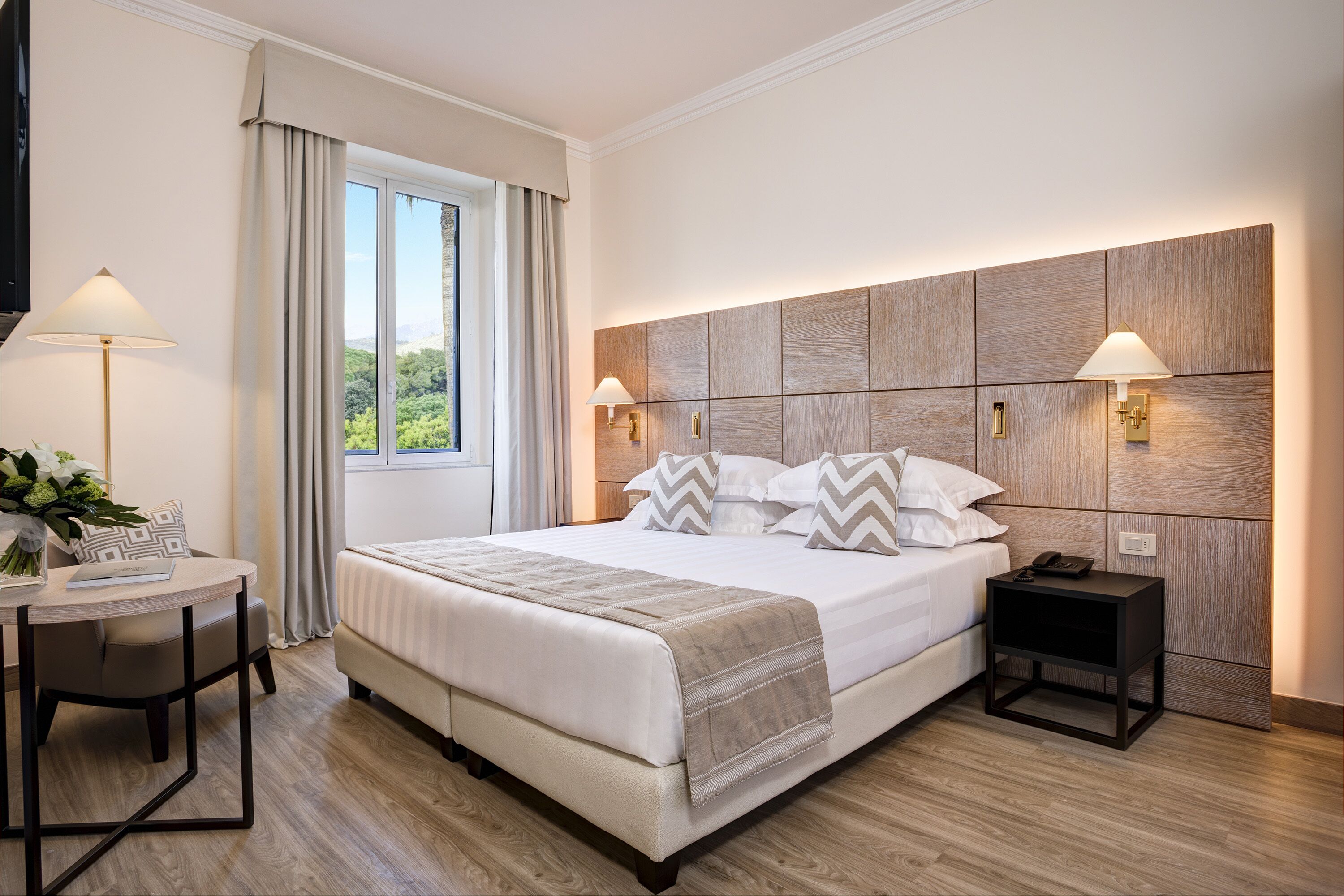 A modern hotel room with a large bed, bedside lamps, a small table with a lamp, and a view of greenery through the window.